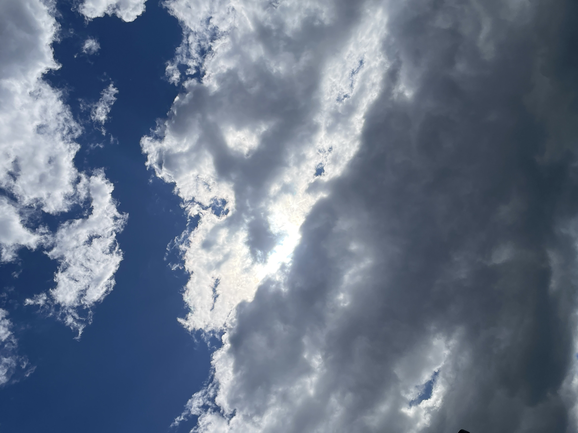 The image shows a view of the sky. There is a large, dark cloud formation occupying much of the right side and upper portion of the image. Parts of the cloud are white where the sun shines through. The left side of the image reveals patches of blue sky with scattered, smaller, white clouds. The location is Unknown, Unknown.