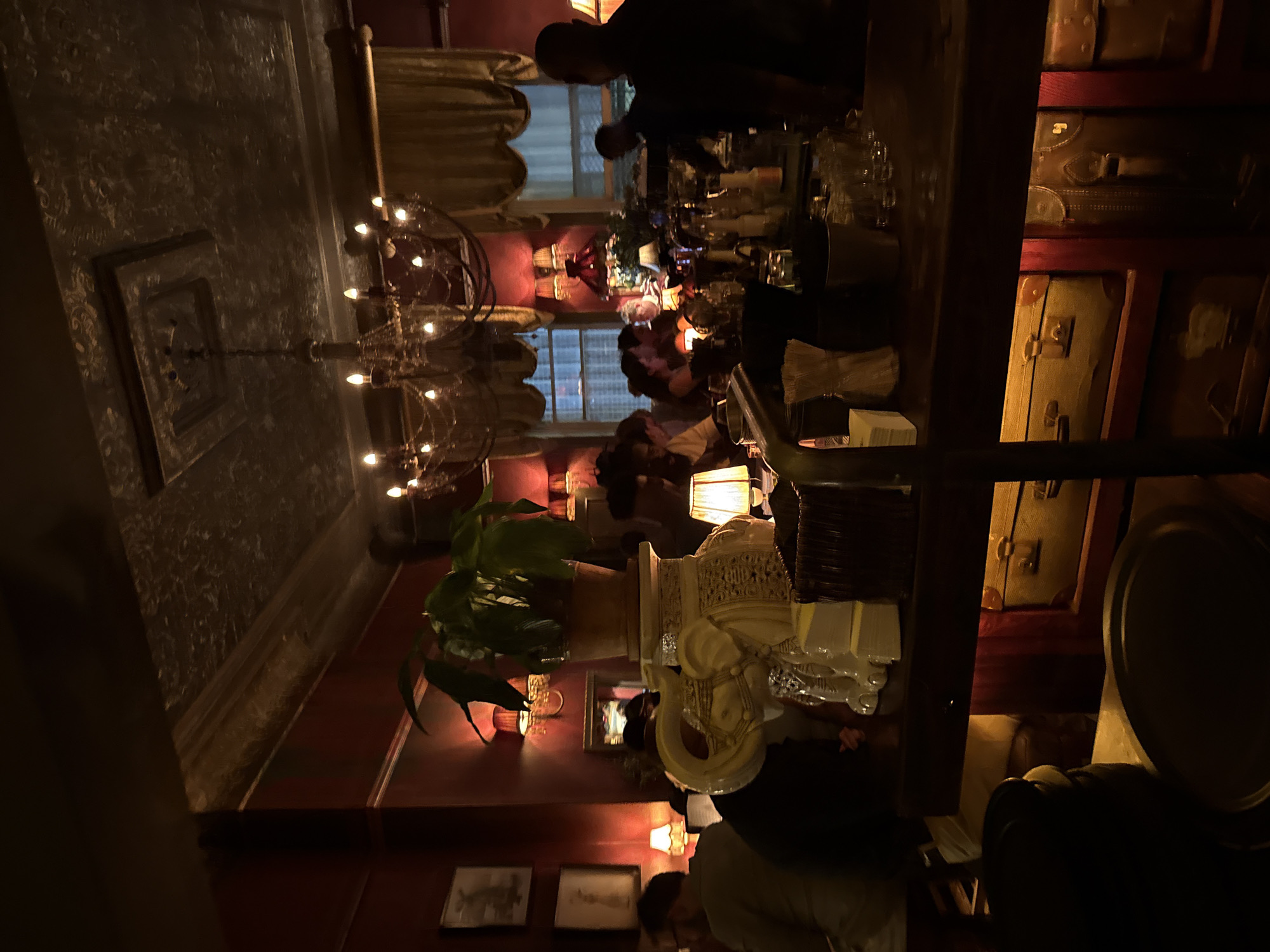 The image captures the dimly lit interior of a restaurant or bar in Cape Town, South Africa. People are seated at a long bar counter. Shelves are stacked with vintage suitcases. The interior design includes ornate chandeliers and wall decor. There is a plant on a table. Patrons are visible, engaged in conversations. A server or bartender is tending to the counter.