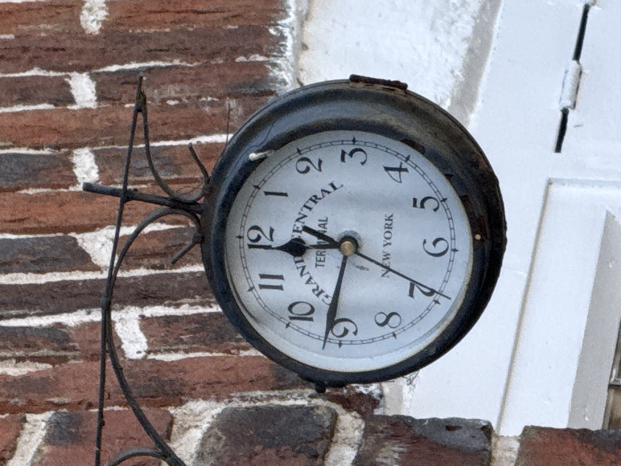 An eye-level close-up shot depicts a station clock mounted on a brick wall. The clock is circular with a black metal frame. Its face is white with black numerals and the words "GRAND CENTRAL TERMINAL NEW YORK" printed on it. The time shown on the clock is approximately 6:23. The brick wall behind the clock is multi-colored with tones of red and brown. A white architectural element is visible to the right of the clock, suggesting it is part of a building facade. The clock's mounting bracket is a dark metal and appears to be attached to the brick wall. The photograph exhibits good lighting, highlighting the details of the clock and its surroundings. The location, Amersham, United Kingdom, is not visibly discernible from the immediate scene but is pertinent information if known through external sources.