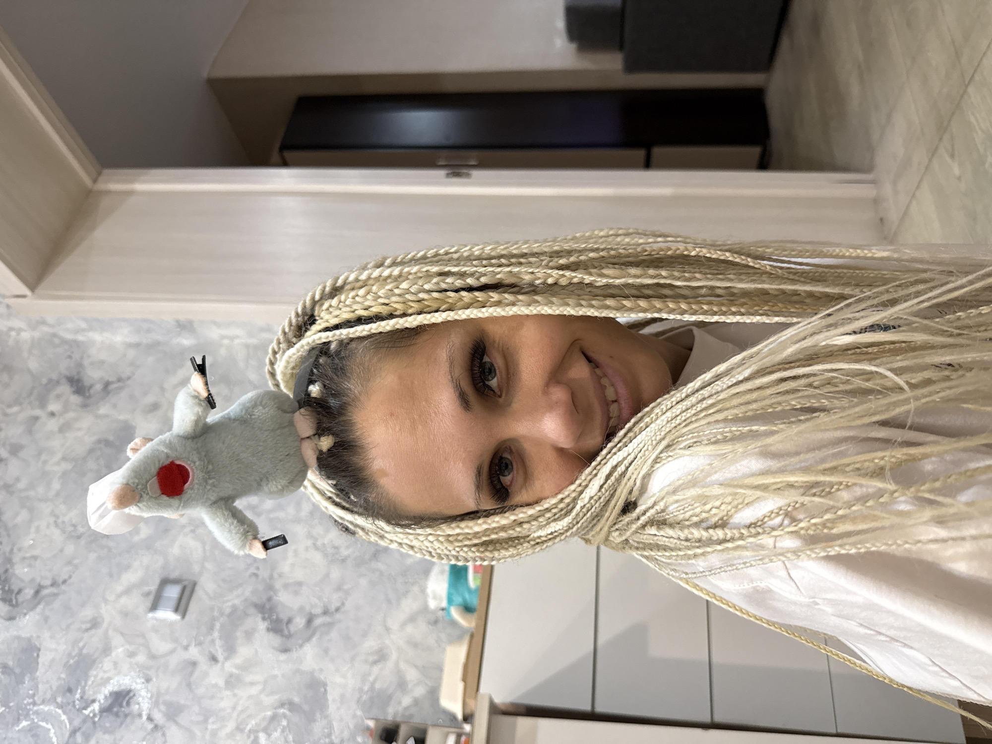 A woman with blonde braided hair is smiling at the camera. A stuffed toy rat wearing a chef's hat is perched on top of her head. The woman is wearing a white t-shirt. The background appears to be an indoor setting, possibly a home, with a gray marbled wall and a doorway leading to another room. There is furniture in the background, including a dresser or cabinet and a tall, dark-colored wardrobe. The floor is tiled. The image does not provide any specific details about the location in the city of Rusanovo, Russia.