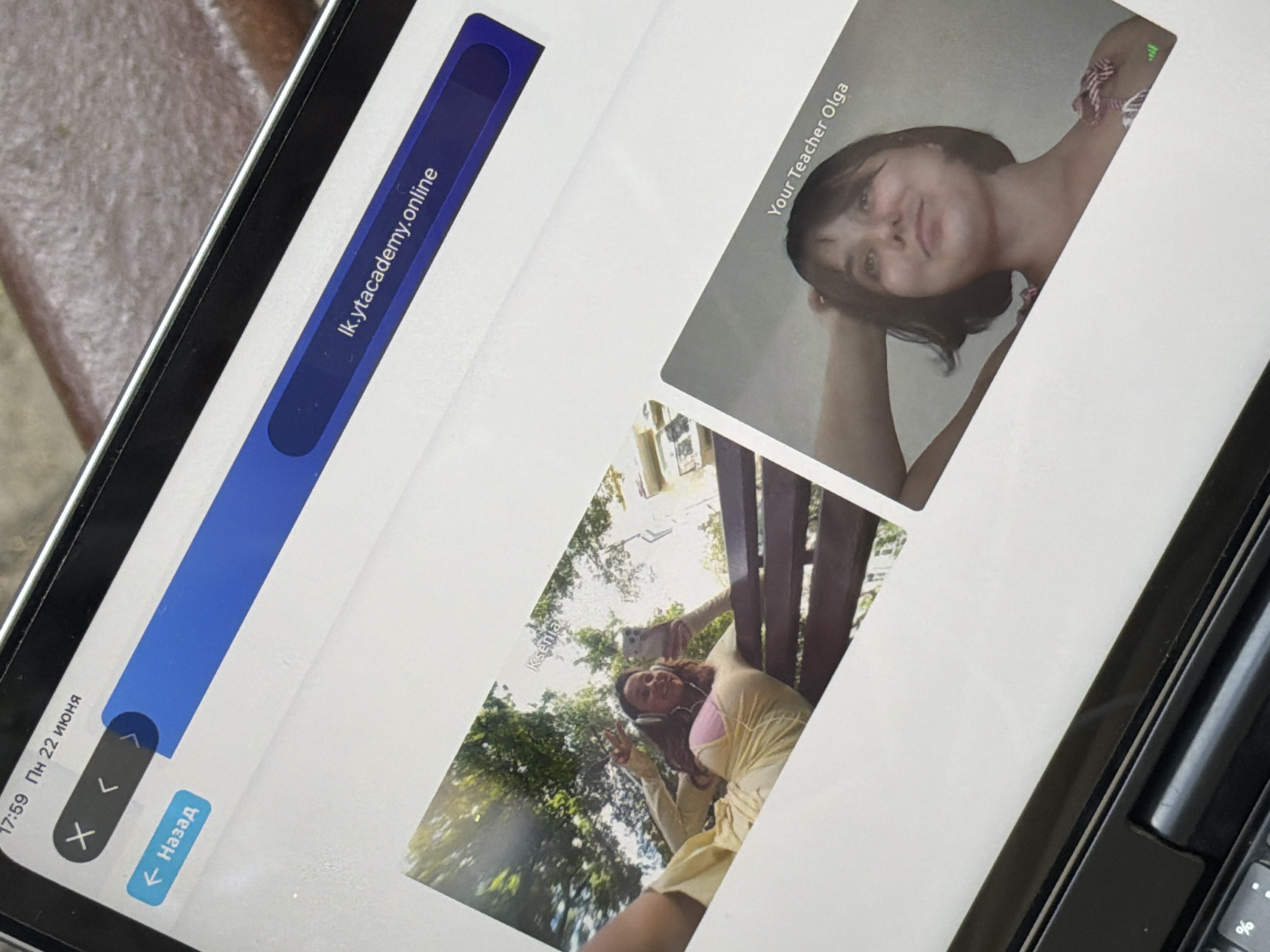 The image displays a digital tablet screen showing a video call interface. On the left, a user named "Ksenia" is visible, taking a selfie with headphones on and a yellow dress. She is sitting on a park bench with trees in the background, suggesting an outdoor setting. On the right, a participant identified as "Your Teacher Olga" is shown. The top of the screen indicates the time is 17:59 on June 22nd and displays a URL "lk.ytacademy.online". The overall context suggests a remote learning or meeting session. The presence of greenery and sunlight implies it is daytime and possibly a warm season.