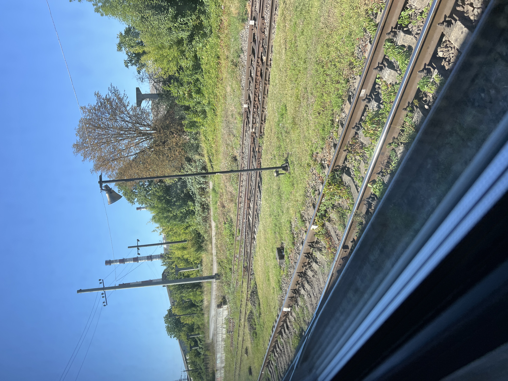 The image depicts a view from a train window of train tracks stretching into the distance under a clear blue sky. Multiple sets of parallel railway tracks are visible, with lush green grass and some patches of dry vegetation growing between and around them. In the background, there are industrial structures, including tall, slender smokestacks with red and white stripes, and utility poles with electrical wires. Dense green trees line the landscape behind the tracks, and a single tree with autumnal foliage stands out against the greenery. A tall, dark pole with a speaker or lamp at the top is positioned near the tracks. The foreground is dominated by the reflection of the sky and some indistinct metallic elements, likely from the train's interior, on the windowpane. The scene appears to be an industrial or semi-industrial area bordering a railway line, with a sense of clear weather and daylight. The location is identified as Ivano-Frankivsk, Ukraine.