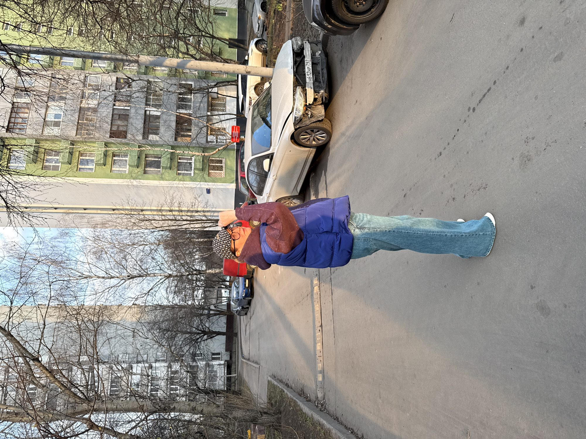 A woman stands on a city street near an accident involving a white BMW with the license plate H612HP797, indicating a Moscow registration. The car is heavily damaged in the front. The street scene appears to be in Moscow, Russia, given the license plate of the car. The woman wears blue jeans, a blue vest, and a brown leather jacket, and she is holding her phone up to her face. A multi-story residential building stands in the background with bare trees.