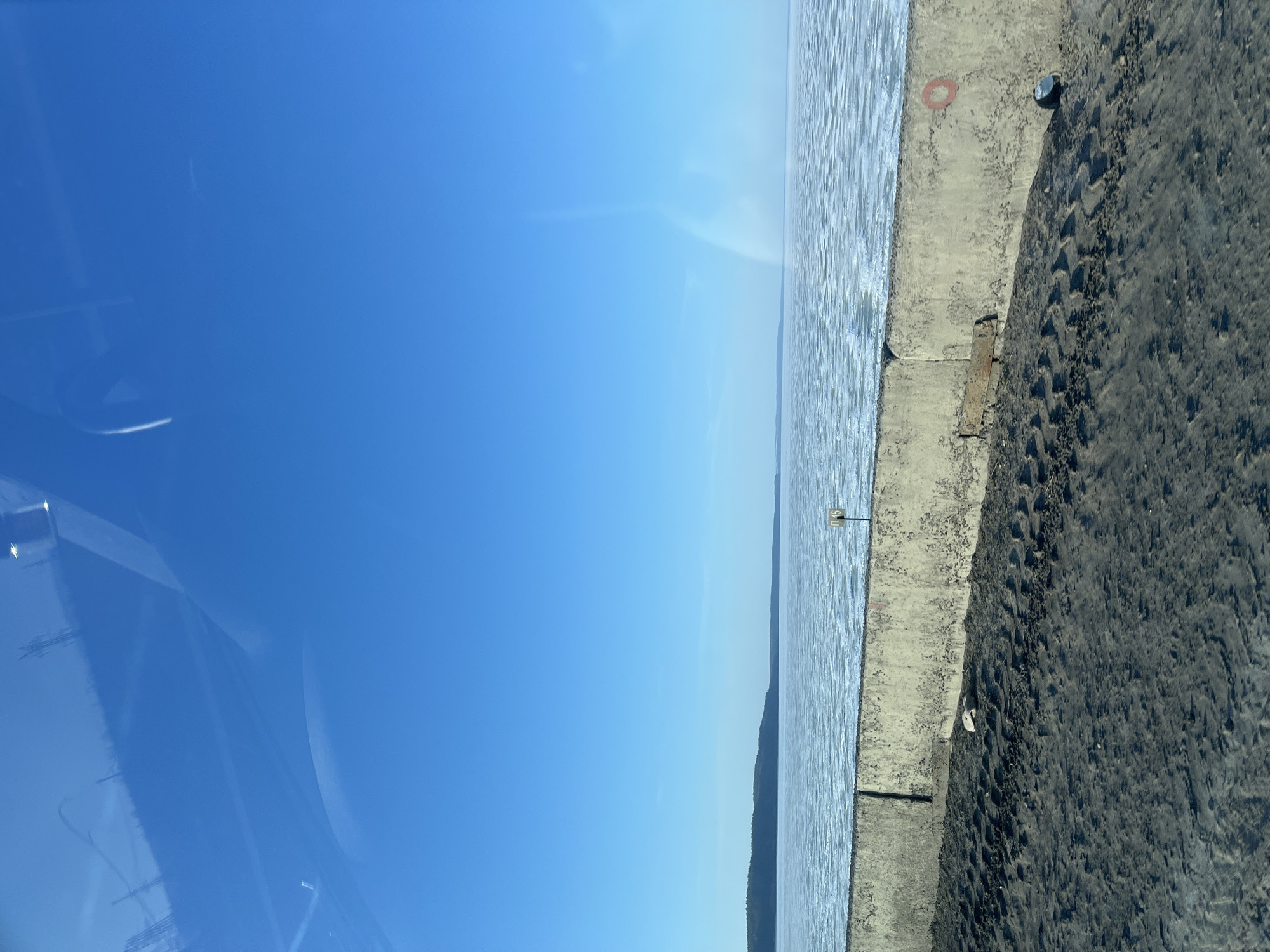 A waterfront view under a clear blue sky. In the distance, a strip of land is visible across a body of water. The foreground features a concrete edge and a dirt surface with tire tracks. The image appears to be taken from inside a vehicle, as evidenced by reflections in the top left. A sign is fixed to the concrete edge. The location is inferred to be in Chaikovsky, Russia. The details of people, subjects, objects, actions, and interactions are not directly observable.