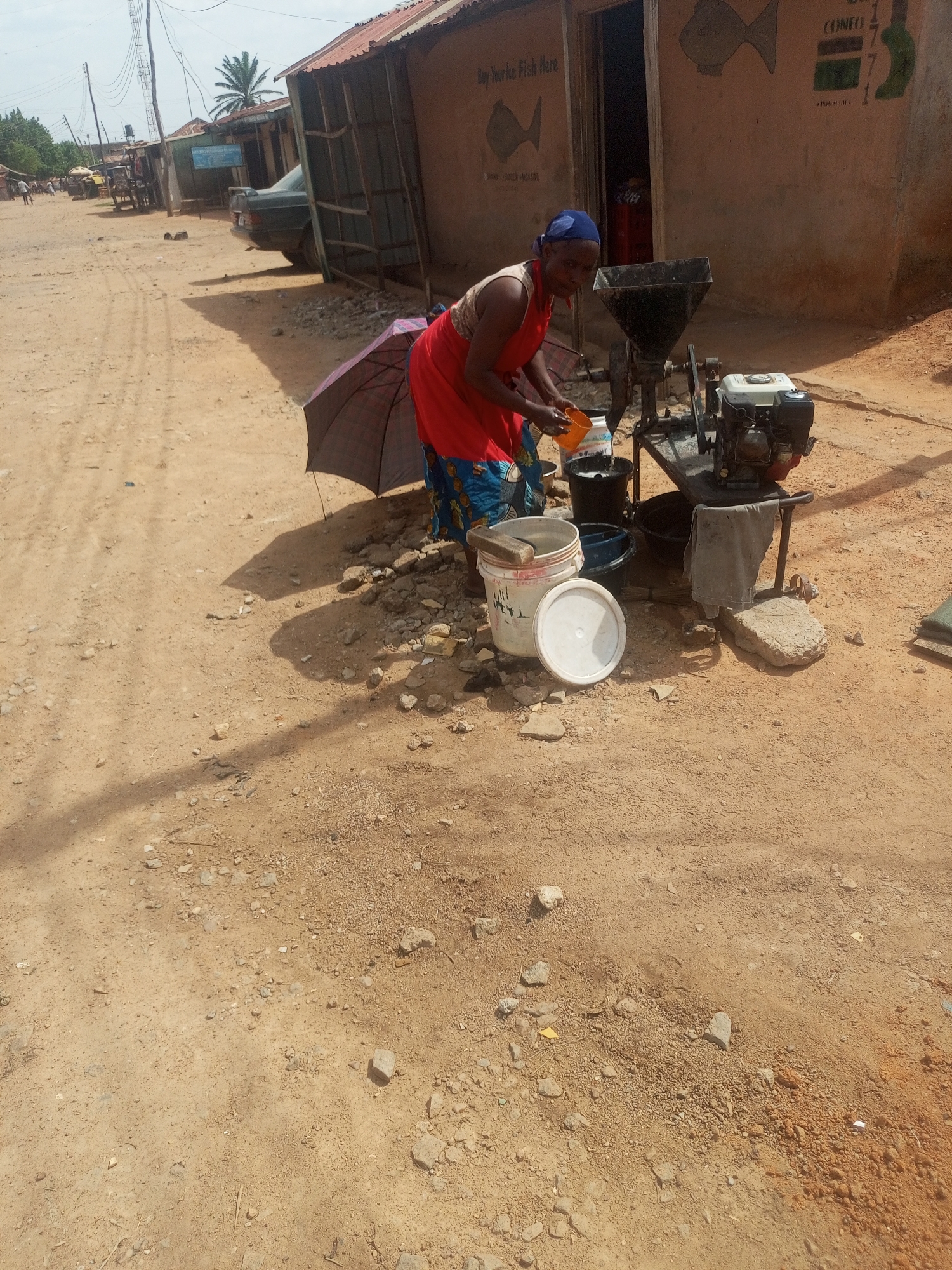 A street scene in Yelwa, Nigeria, shows a woman in a red apron and blue headscarf operating a small, gas-powered milling machine. She is pouring liquid from an orange cup into the machine. Several buckets are nearby, along with an open umbrella and a white bucket with its lid askew. The woman is positioned in front of a building with painted images of fish and the word "CONGO." The ground is dirt with scattered rocks. A dirt road runs along the left side of the image, with other buildings and vehicles visible in the distance. Overhead power lines are present.