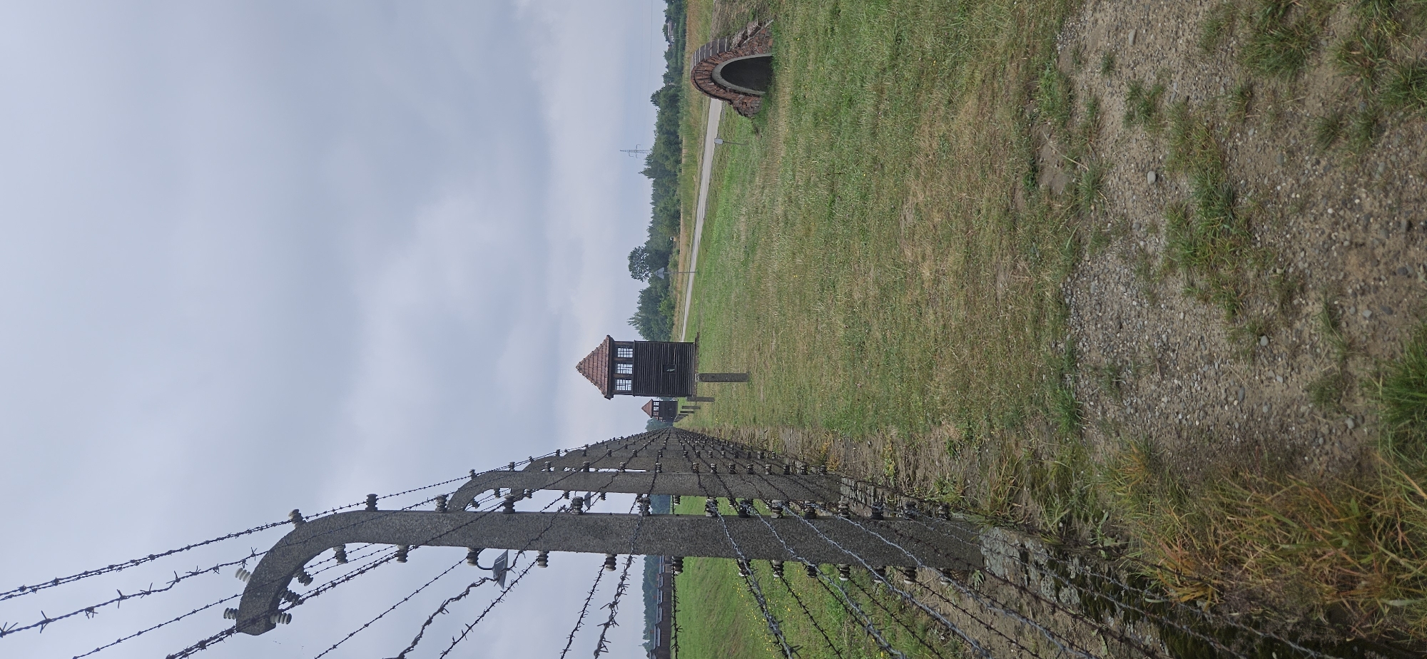 The image depicts a view from within the grounds of Auschwitz-Birkenau Memorial and Museum, located in Brzezinka, Poland. In the foreground, a weathered barbed wire fence runs along a gravel path, with concrete posts topped with insulators supporting the wire. Beyond the fence, a grassy field stretches towards the horizon. Two wooden watchtowers with red-tiled roofs stand in the midground, positioned along what appears to be an internal road or pathway. Further into the distance, to the left, a row of low-lying wooden structures, possibly barracks, are visible. To the right of the watchtowers, a small arched structure made of brick is partially visible, set into the ground. The sky above is overcast with gray clouds. The overall scene conveys a somber and historical atmosphere, characteristic of the former concentration camp.