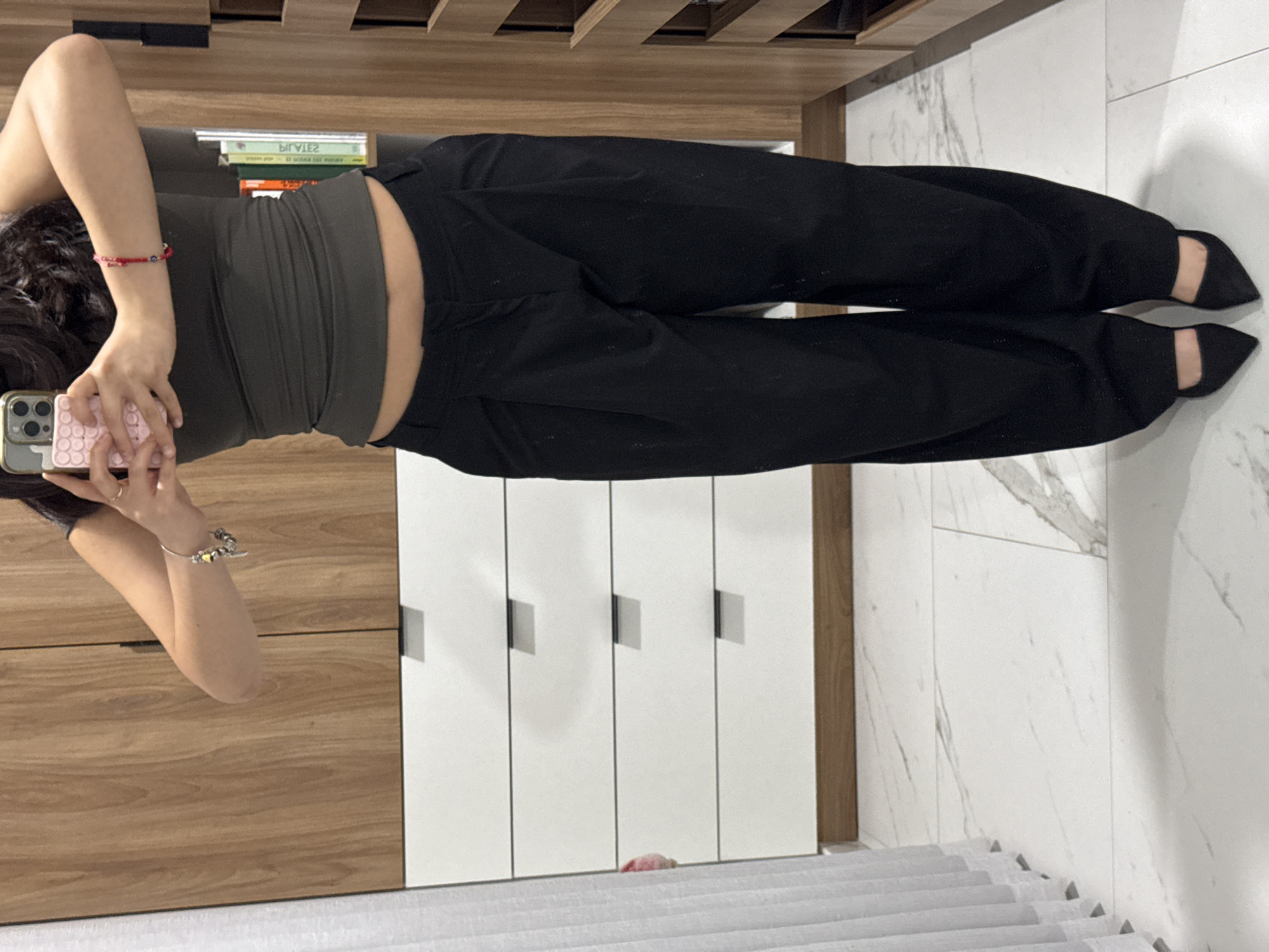 A person is depicted from the upper chest to the feet, taking a mirror self-portrait indoors. The subject holds a smartphone with a pink textured case, obscuring their face. They are wearing a dark olive green sleeveless crop top that reveals a portion of the abdomen, black pleated wide-leg trousers, and black pointed-toe heels. A silver charm bracelet is on the subject's left wrist, and a red beaded bracelet with a blue charm is on the right wrist. The background features a modern interior with reflective surfaces. To the left, a white pleated curtain is partially visible next to a white five-drawer dresser with dark handles and light wood trim. The central and right background consists of light wood-grain wardrobe doors, which reflect elements of the room. A shelving unit on the far right contains books, one of which appears to be titled "PILATES" when viewed in its reflected form. The floor is covered with white marble-patterned tiles. There are no visual elements within the image to determine its location in the city of Puebla, Mexico.