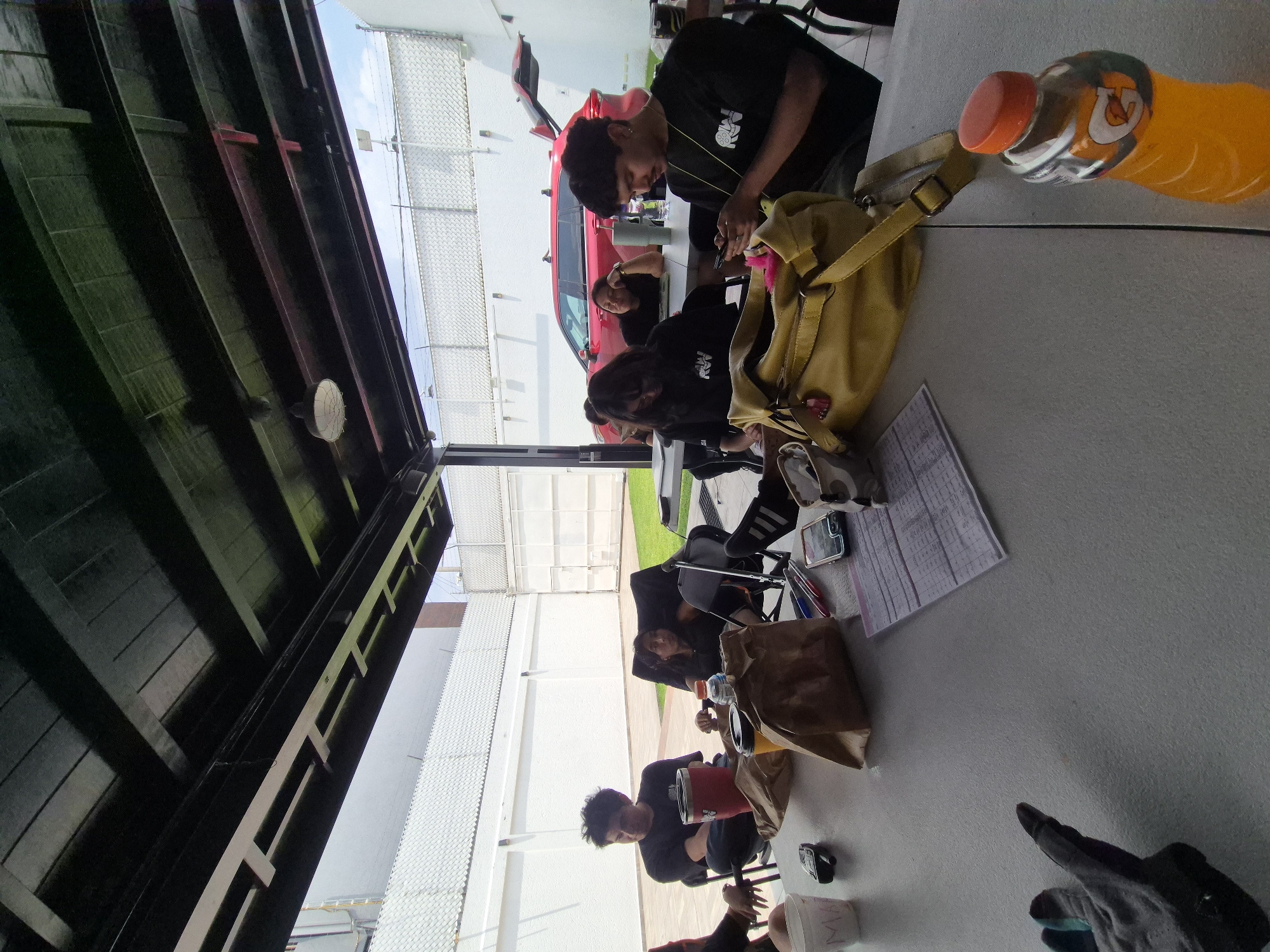 A photograph taken in Puebla, Mexico, depicts a group of people sitting at a table, seemingly engaged in various activities. The location appears to be outdoors under a covered structure, potentially a patio or open-air dining area.
Several individuals are visible, most are sitting at the table wearing black clothing and they all appear to be concentrated on something on the tabletop. There is a red car visible.
Objects on the table include a yellow bag, a partially consumed bottle of Gatorade, papers, and a variety of personal items, such as phones.