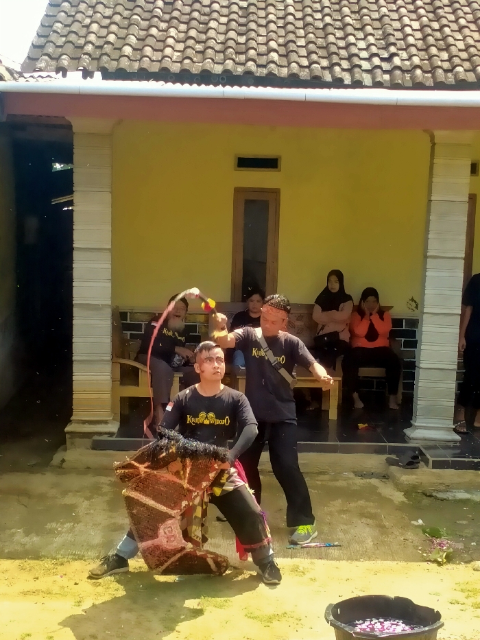 The image captures an outdoor ritual or performance in Tawangsari, Indonesia. In the foreground, a male participant, splattered with a reddish substance on his face and upper body, is positioned in a low, wide stance, holding or integrated with a large, damaged, dark brown and red horse-shaped prop. He wears a black long-sleeved shirt with an Indonesian flag emblem and text. Immediately behind him, a second male participant, similarly splattered, wields a whip-like object with a yellow and red handle over the first man. A third male participant, partially obscured to the left, also appears to be involved with a whip, creating a visible spray of reddish droplets. All three active male participants wear black shirts. In the background, on the tiled porch of a yellow-walled house with a brown tiled roof and white supporting pillars, three women are seated on a wooden bench observing the event. A black bucket containing water and flower petals is situated in the lower right foreground. The ground in the immediate performance area is unpaved, appearing as dirt or worn concrete.
