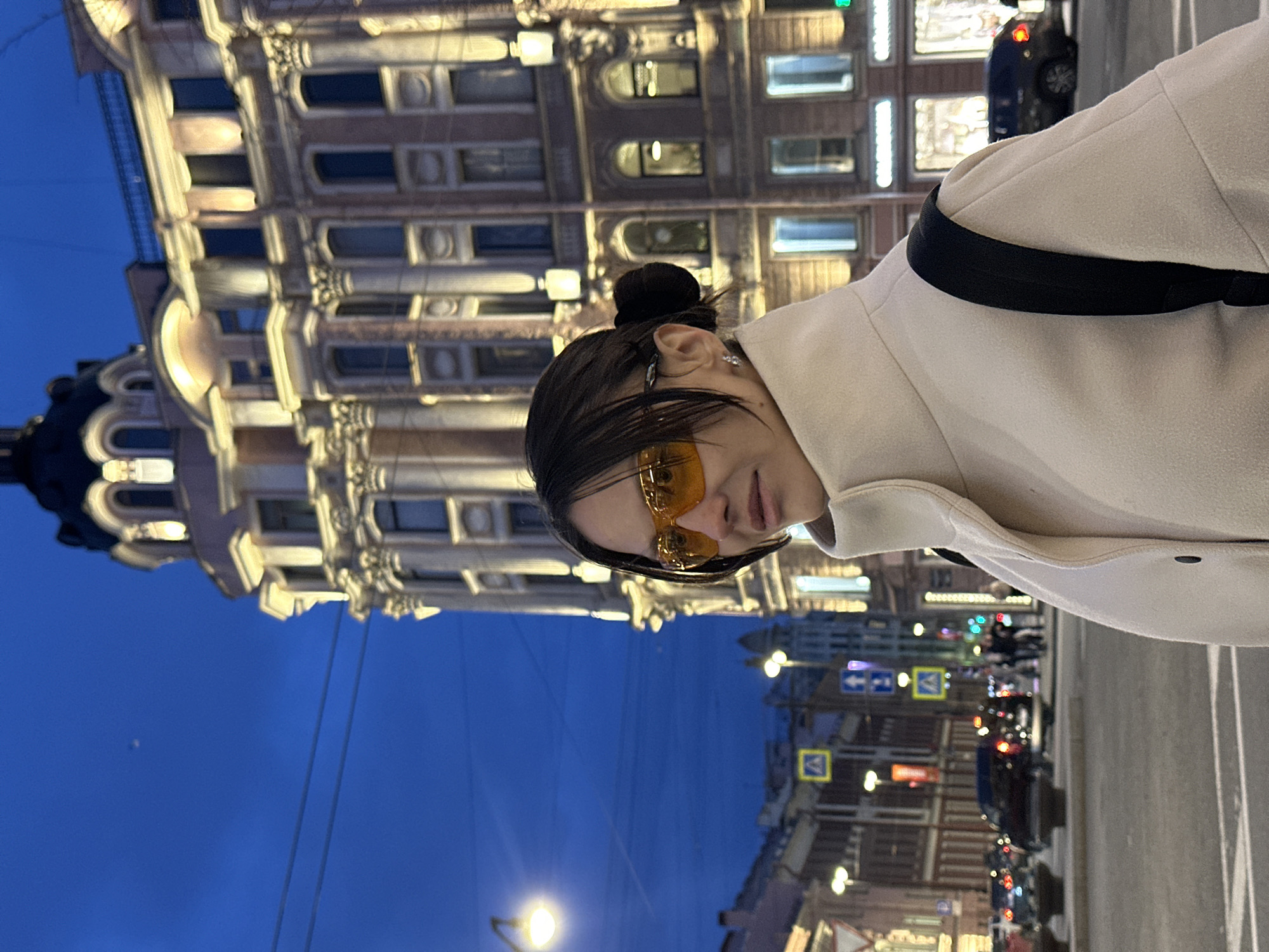 The image, when rotated to its correct orientation, captures an evening or nighttime street scene centered around a young woman. She is positioned towards the right of the frame, looking slightly downwards and to her right, wearing distinct orange-tinted sunglasses. Her dark hair is styled in a bun on top of her head. She is dressed in a light-colored, possibly beige or off-white, coat, with a prominent black strap crossing her left shoulder, potentially from a bag. A small, sparkling earring is visible in her right ear.
The background features a magnificent, multi-storied building on the left, characteristic of European architecture, likely in Saint Petersburg. It boasts elaborate architectural details, including ornate windows, decorative cornices, and pilasters, all illuminated by warm interior lights that glow against the deep blue twilight sky.
To the right and further into the background, a city street unfolds, bustling with evening activity. The red taillights and white headlights of multiple vehicles are visible, indicating traffic. Several streetlights cast bright circles of light onto the road. Various traffic signs, including a blue rectangular sign with white arrows and a yellow square sign with a black pedestrian crossing symbol, are visible along the street. The overall impression is one of a vibrant urban setting during the early night hours, combining architectural grandeur with city life. No clear text is legible within the frame.