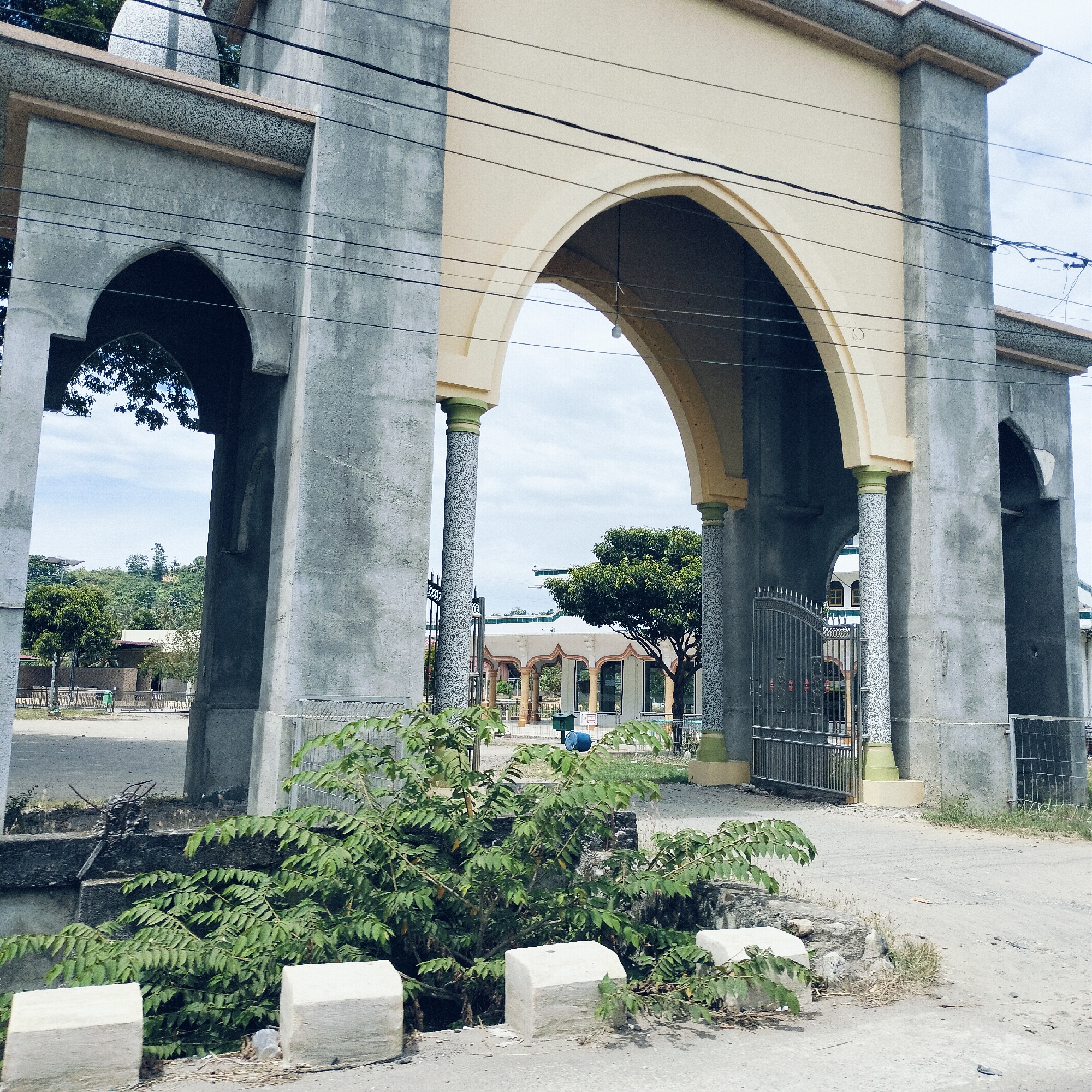 A wide exterior shot captures a large, ornate arched gateway, likely the entrance to a mosque or religious complex, in Keudealuerheung, Indonesia. The gateway is constructed from rough-textured grey concrete, with the inner surfaces of the main arch and some upper sections painted a light beige. Its columns feature a speckled, granite-like finish and light green bases.
Through the prominent central arch, a paved courtyard or road leads to other buildings with matching architectural styles, including arched openings and supporting columns. A black metal gate with decorative elements is visible beneath one of the arches on the right. In the distance, beyond these structures, hills covered with lush green trees are visible under a bright, overcast sky. A small dome, typical of mosque architecture, can be seen at the top left, peeking above the gateway's structure.
In the immediate foreground, a vibrant green leafy bush grows beside the road, partially obscuring white concrete blocks lining the edge of what appears to be a drainage ditch. Power lines stretch across the upper portion of the frame, crisscrossing the gateway. A blue barrel and a green, rectangular container are situated in the courtyard beyond the arch. The scene is static, devoid of people or discernible activity, captured during daylight hours under a bright sky. No visible text is present.