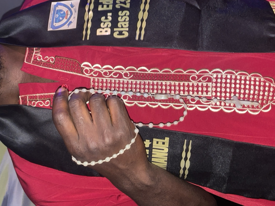 A person is seen wearing a red shirt with decorative trim, and a black graduation stole embroidered with “BSc. Ed.” and “Class 23.” The stole features a college emblem. The person's hand holds a rosary. A book titled "+ MANUEL" is adjacent to the graduation sash.