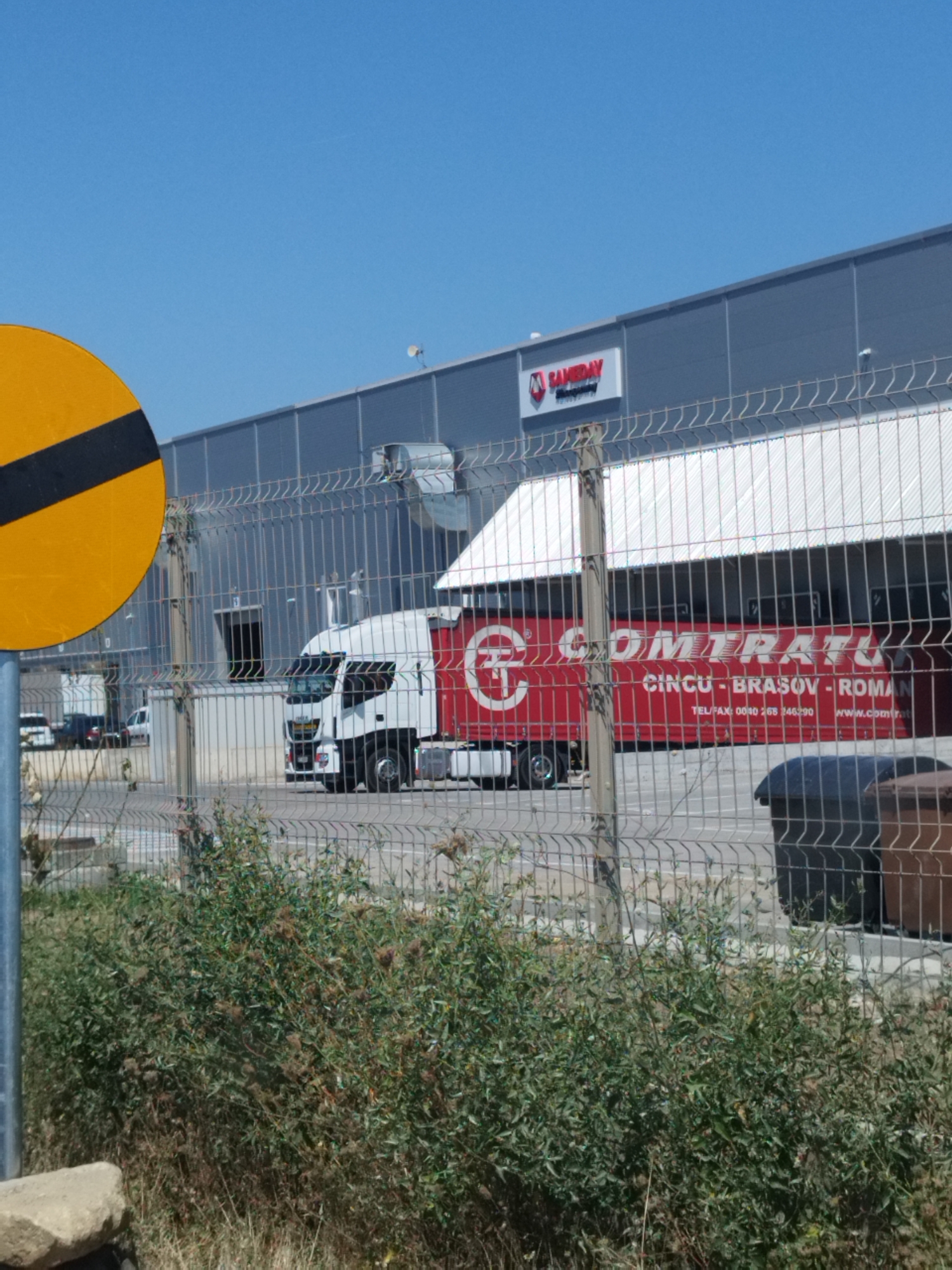 The image captures a sunny outdoor scene in Bragadiru, Romania, likely near a commercial or industrial area. In the foreground, a chain-link fence runs across the width of the frame, with overgrown green foliage and some dry grass visible below it. To the left, a large yellow circular sign with a black diagonal stripe is partially visible.

Behind the fence, a white truck with a red trailer is parked, its rear end facing the viewer. The trailer is branded with red lettering that reads "COMTRATU," with "CINCUI - BRASOV - ROMAN" written below it. Telephone and website information are also present on the trailer in smaller print. The truck appears to be in front of a large, modern grey building with a white, sloped roof. A loading bay or dock is visible on the building. Further back, other vehicles, including cars and vans, are parked in front of the building, suggesting a functional logistics or distribution hub. The sky is clear and blue.