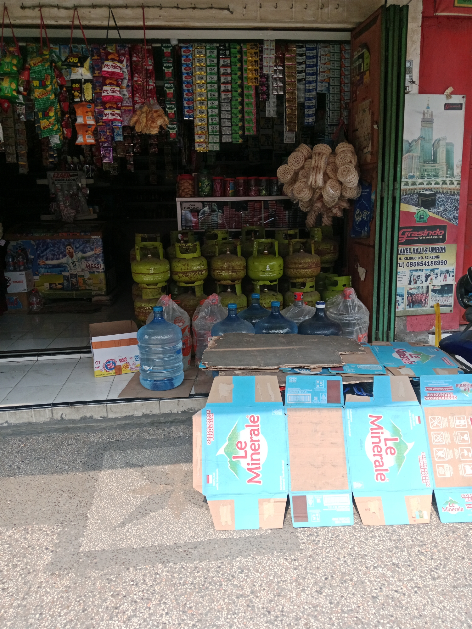 A street-level view captures an outdoor storefront in Kediri City, Indonesia. The scene is dominated by an array of goods displayed for sale. In the foreground, several blue cardboard boxes with the "Le Minerale" logo are arranged, some open and others closed, suggesting a retail or delivery operation. Behind these, a row of large blue water jugs are visible. Further back, stacked green propane gas tanks are lined up, indicating a fuel supply business. The storefront itself is filled with shelves stocked with various packaged goods, including small bags of snacks and drinks hanging from the ceiling. A poster advertising "Grasindo Travel Haji & Umroh" is prominently displayed on the right side of the frame, including contact information. The environment appears to be a typical Indonesian urban street setting, with a paved surface in front of the store and a building facade forming the backdrop. The lighting suggests it is daytime. There are no people clearly visible in the image.