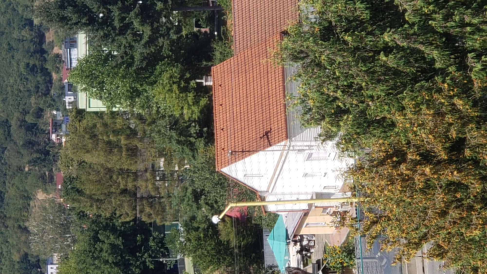 The image shows a view from an elevated position, overlooking a residential area nestled on a hillside. In the foreground, there are two houses, one with a white facade and a prominent orange-tiled roof, and another smaller, partially visible house with a light yellow facade. A tall yellow lamppost is visible in front of the white house. Trees with green and yellow foliage are abundant throughout the scene. In the background, several other houses and buildings are visible, along with a dense forest covering the hillside. The sky is clear and bright, suggesting a sunny day. The overall impression is of a quiet, suburban neighborhood in a hilly region. Based on the architectural style and the presence of a European lamppost, this scene is likely located in Prešov, Slovakia, a city known for its hilly terrain and mixed architectural styles.