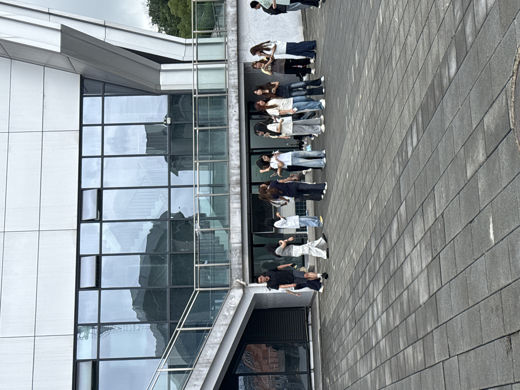 The image captures a group of young adults standing and walking on a paved plaza in front of a modern building. The building features large glass windows that reflect the sky and surrounding environment, along with a prominent architectural design with angular elements. The plaza is paved with dark gray rectangular tiles.
In the foreground, several individuals are visible. A man in a black polo shirt and dark shorts walks from left to right, looking forward. To his right, a woman in a light gray t-shirt and white pants walks in the same direction. Further to the right, a group of young women are standing, some looking at their phones. One woman in a white t-shirt and blue jeans is seen from the back, facing away from the camera. Another woman in a gray and white striped t-shirt and black pants is interacting with a woman in a white t-shirt and blue jeans. A man in a dark shirt and light shorts is visible on the far right, partially obscured.
The background shows the building's facade with rows of windows and a staircase visible through the glass on the upper levels. The overall scene suggests a casual gathering or break time for students or young professionals in an urban or institutional setting. The location is identified as Jiangjin City, China.