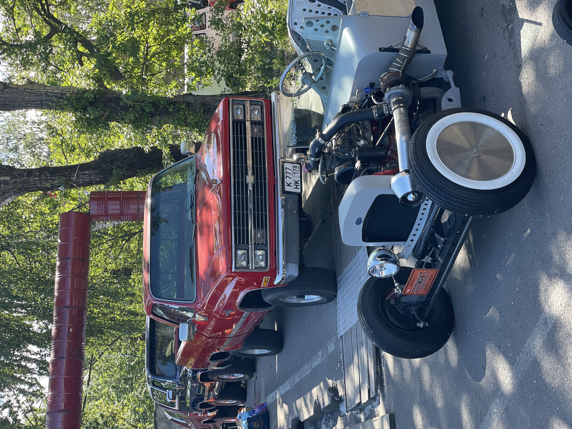 This is a daylight, eye-level, medium shot of several vehicles parked outdoors. In the foreground, a light gray, partially assembled hot rod with a visible engine and turbocharger is parked on asphalt. The hot rod has a single headlight and a license plate that reads "FROMRKT". Behind the hot rod, a red Chevrolet pickup truck is parked. The truck has a visible license plate that reads "K 777 HK 178". Further back, a dark-colored pickup truck is visible, followed by another vehicle. The vehicles are parked in a row on a surface that transitions from wooden planks to asphalt. In the background, lush green trees and foliage are abundant, suggesting a park or a residential area. A red pipe structure is visible overhead, stretching across the frame. The scene appears to be set in Saint Petersburg, Russia, indicated by the vehicle license plate format.