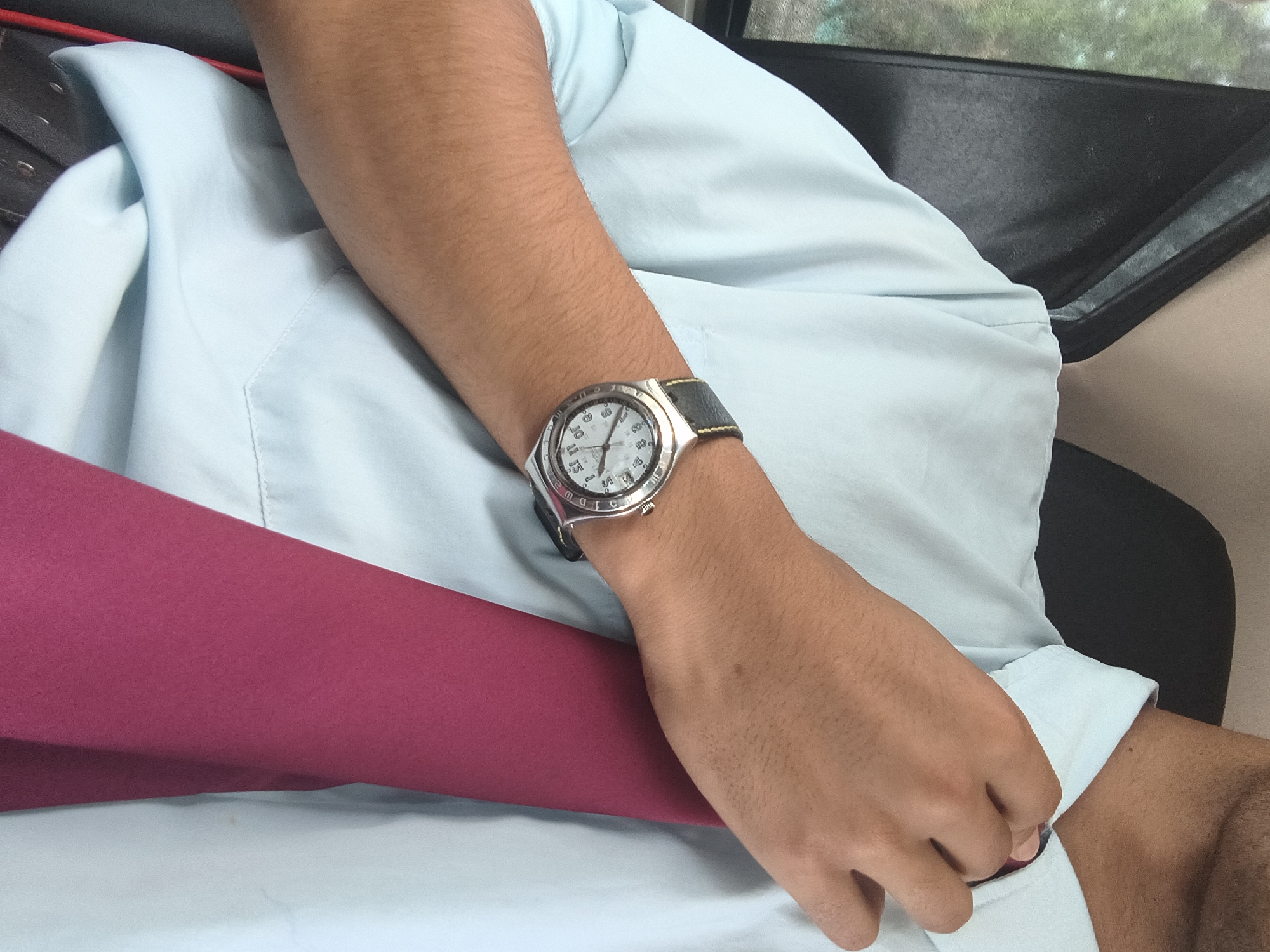 The image shows a close-up of a person's arm resting on a light blue shirt. The person is wearing a silver watch with a dark strap on their left wrist. The watch face has Roman numerals and a date display. A portion of a dark seat is visible in the upper right corner, and a pink fabric is draped across the lower left portion of the frame. The scene appears to be indoors, possibly in a vehicle or a waiting area. The lighting suggests it is daytime. There is no visible text in the image.