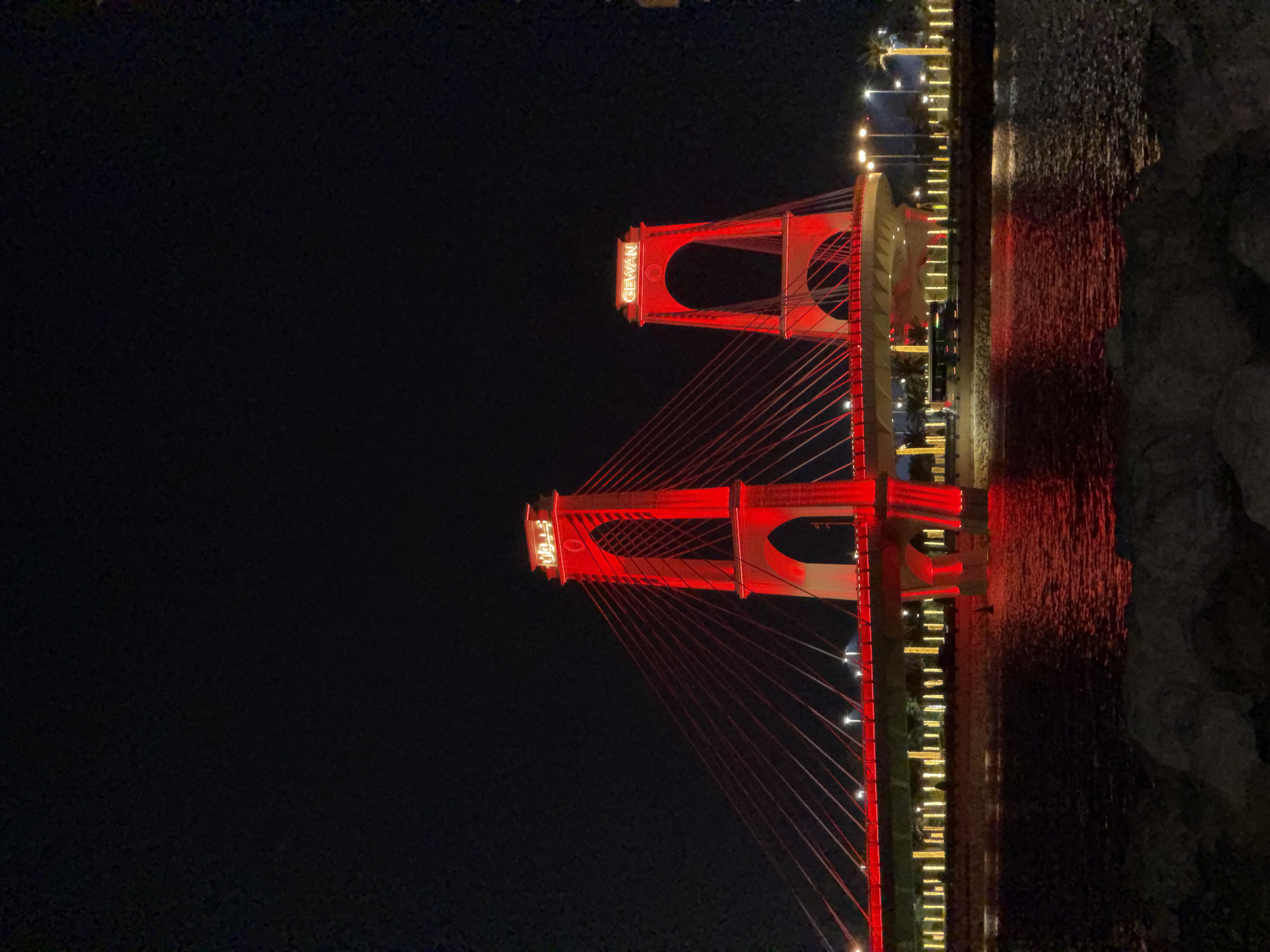 A suspension bridge is illuminated in red at night. The bridge has two tall, arch-shaped towers, each with the word "GEWAN" lit up in red on top. The cables of the bridge are also red. In the background, there are streetlights and palm trees. The water in the foreground reflects the red lights of the bridge. There are no people visible in the image.