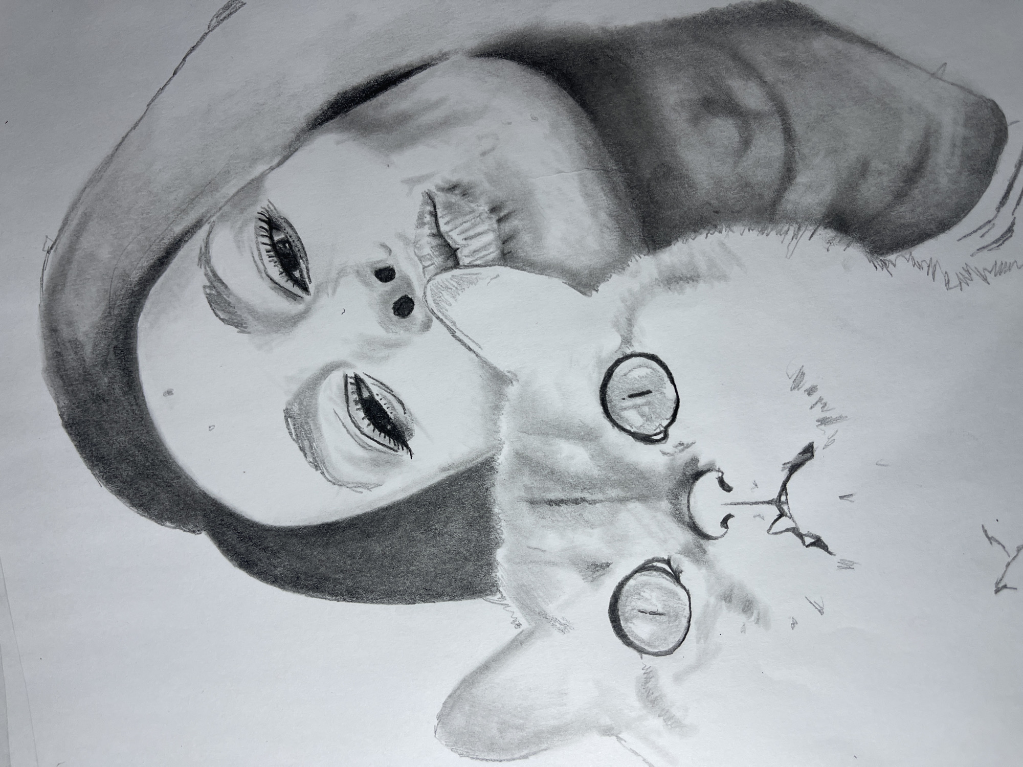 This is a monochromatic pencil drawing rendered on white paper, depicting a close-up portrait of a woman and a domestic cat. The woman is positioned in the upper right portion of the composition, her head slightly tilted. Her visible right eye features defined lashes, and her lips are pursed in a "kissing" expression. Her right hand's fingers are seen near her chin. Dark shading indicates her hair framing her face and extending over her shoulder. The cat occupies the lower-left and central area, its head facing directly forward. It has large, round eyes with thin vertical pupils, a discernible nose, and a slightly open mouth revealing two small lower fangs. Its fur is rendered with varying shades of gray to suggest texture. The woman and cat are in close proximity, implying an affectionate interaction. The background is an undetailed white, indicating no specific environment within the drawing itself. The location of this artwork is the city of Jos, Nigeria.