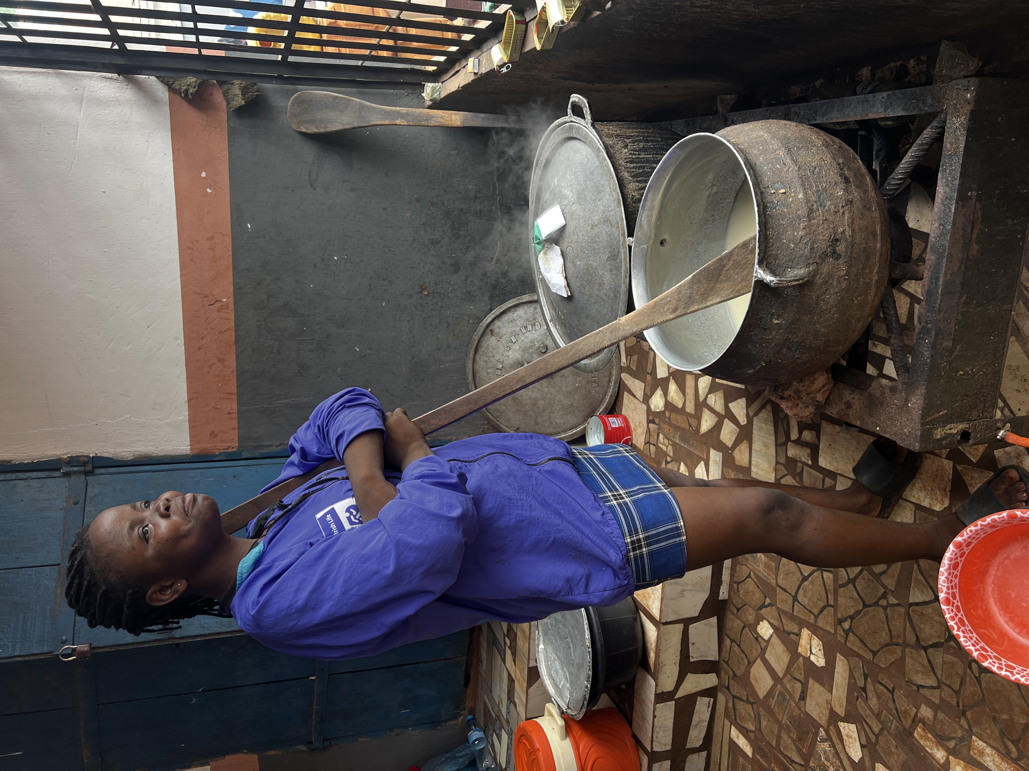In Nkpor, Nigeria, a woman stands by a cooking station outdoors, her arms crossed over her chest. She wears a blue jacket with an "Irish Life" logo and a blue and white plaid skirt. She is facing to the right, looking off-camera. In front of her, a large, dark pot is on a metal stove, emitting steam. Several other large metal pots with lids are also visible in the background. The ground is paved with irregularly shaped tiles. A blue door with a lock is behind the woman. The scene suggests food preparation is underway in an open-air setting.