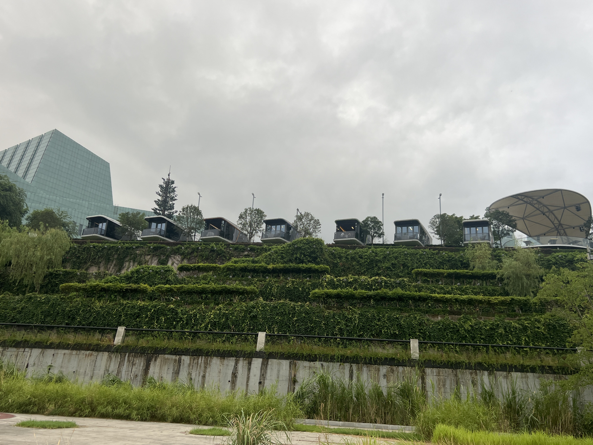 The image depicts a terraced landscape in Jiangbeicheng Subdistrict, China, characterized by lush greenery and modern architecture. A series of seven identical, elevated pods are situated along the top tier of the terraced hill, each with large windows and balconies, overlooking the scene. The hill itself is covered in dense green vegetation, creating a cascading effect with visible horizontal layers.
To the left, a striking, angular glass building with a reflective facade dominates the skyline. To the right, a large, curved, white canopy structure, resembling an amphitheater or a modern pavilion, is partially visible. The foreground shows a concrete retaining wall with vegetation growing on top, and further down, a paved area with tall green grass. The sky is overcast with a thick layer of gray clouds. No people are present in the image.