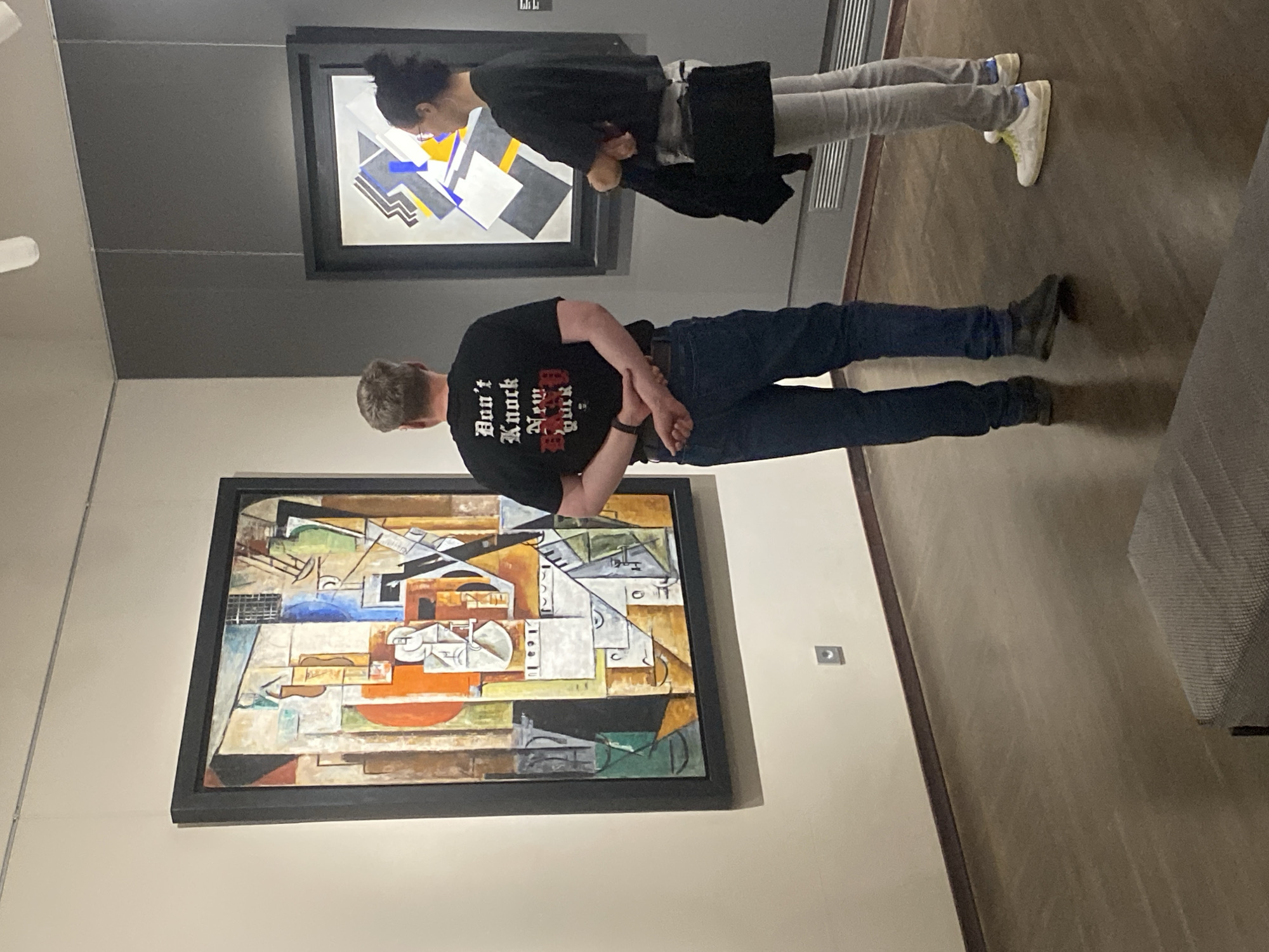 Two individuals are observed in a museum gallery in Yekaterinburg, Russia, standing before abstract paintings. The male, positioned on the left, wears a black t-shirt with gothic-style lettering that reads "Don't Knock New York" and dark blue jeans. He has his arms clasped behind his back. The female, on the right, wears a black top, light grey jeans, and white sneakers with yellow laces. She is holding a dark jacket over her left arm and has her arms crossed.
The gallery walls are painted in neutral tones, with a prominent light beige wall on the left and a darker grey wall on the right. Two framed abstract artworks are displayed. The larger painting on the left features a composition of geometric shapes and vibrant colors, including orange, blue, green, and white. The smaller painting on the right is predominantly monochromatic with accents of blue and yellow. The floor is made of dark wood. A light fixture is visible on the ceiling above the paintings. A small electrical outlet is mounted on the beige wall. In the foreground, a dark grey upholstered bench or seating area is partially visible. The overall environment suggests a cultural institution dedicated to art exhibition.