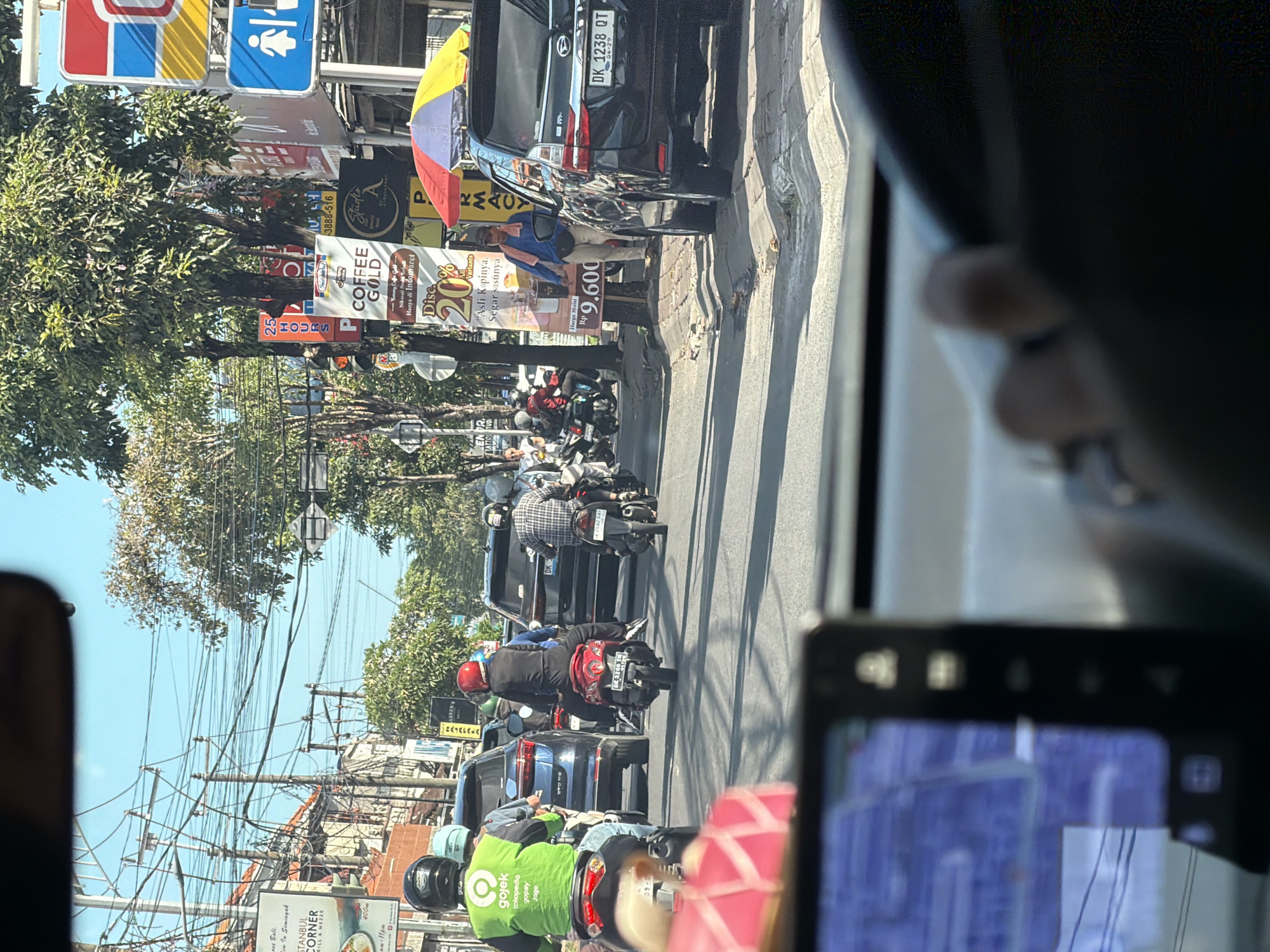 The image captures a street scene in North Kuta, Indonesia, observed from inside a vehicle. The traffic consists of cars and numerous motorcycles, some driven by Gojek riders in green jackets. Street signs and banners advertise local businesses, including "Istanbul Corner" and "Coffee Gold," promoting Indomaret discounts. A black car is parked along the roadside, and a person appears to be standing next to it. A road sign indicating the presence of restrooms. The sky is blue, partially obscured by trees and a network of overhead power lines. A navigation screen is visible in the foreground within the observer's vehicle.