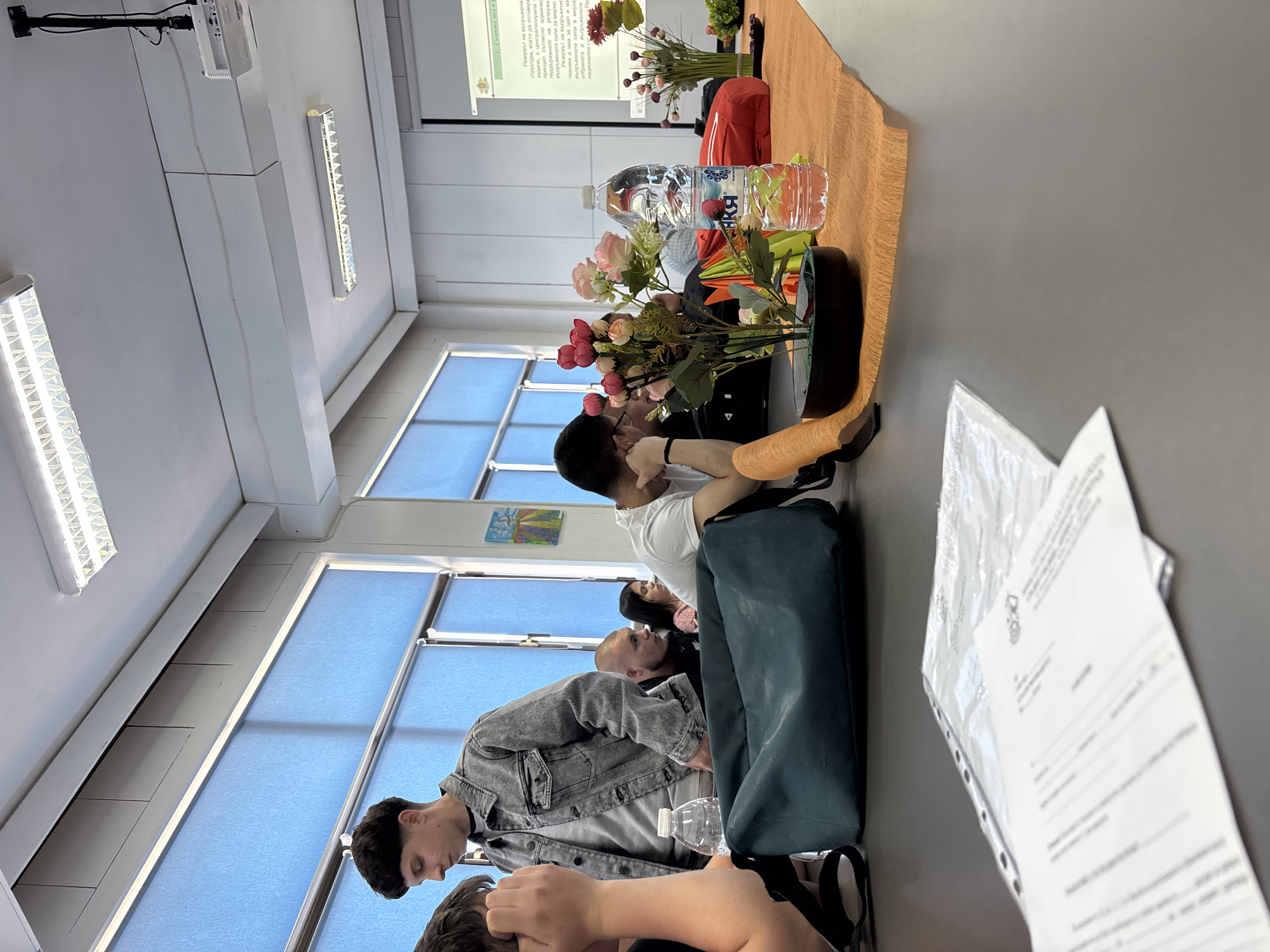 A classroom setting is captured in this image, likely in Zlatograd, Bulgaria. Several individuals are seated at a table, seemingly attending a presentation or lecture. In the foreground, papers are stacked. On the table are arrangements of flowers, both artificial and real, along with water bottles. A projector screen displays text in Cyrillic. Fluorescent lights are visible on the ceiling, and light comes in through windows with blue blinds.