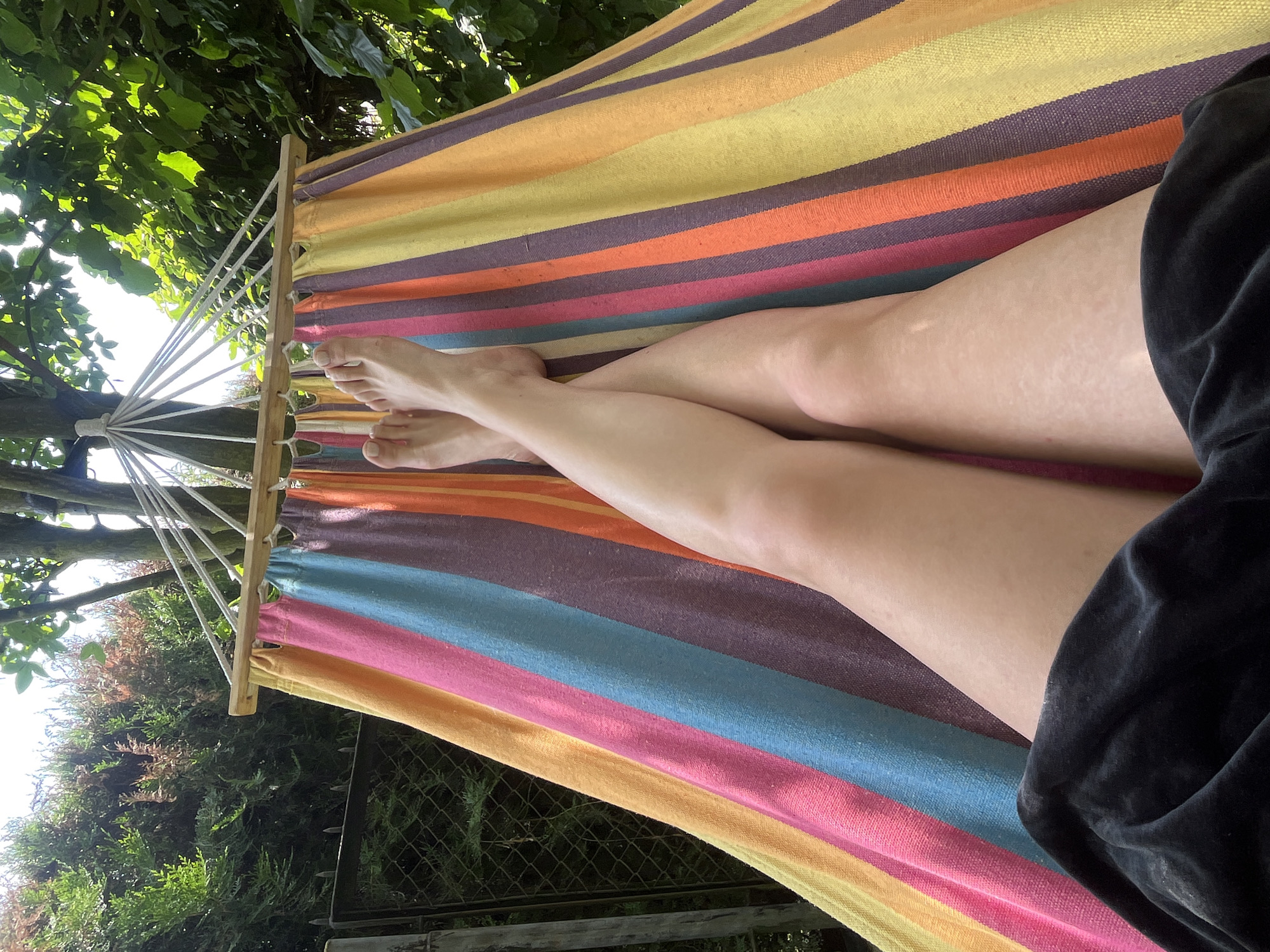 The image shows a person lying in a hammock outdoors. The hammock has multi-colored stripes in shades of yellow, orange, pink, blue, and purple. The person's legs and feet are visible, crossed at the ankles. They are wearing dark clothing at the bottom of the frame. The background consists of green trees and foliage, suggesting a natural setting. There is also a glimpse of a fence and some bushes on the left side of the image. The lighting indicates it is daytime, possibly sunny, with dappled light filtering through the leaves. There is no indication of the location being in Krakow, Poland.