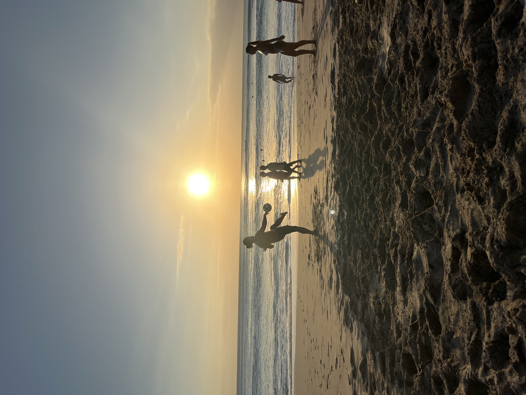 This image captures a serene sunset scene on the beach in Kuta Selatan, Indonesia. Silhouettes of people are scattered across the sandy shore and in the shallow water. In the foreground, a person is captured mid-action, playing with a ball on the sand, with their body angled as if kicking it. Further back, a group of three individuals walks near the water's edge, and another person stands slightly offshore. A woman in a bikini is prominently positioned on the right side of the frame, facing left. The sun, a brilliant orb, hangs low in the sky, casting a warm golden glow that reflects off the gentle waves of the ocean. The sky transitions from a pale blue at the top to a soft orange and yellow hue closer to the horizon, with a hint of a cloud. The sand in the foreground is textured with footprints and undulations, creating a natural foreground element. The overall atmosphere is one of leisure and tranquility as the day ends.