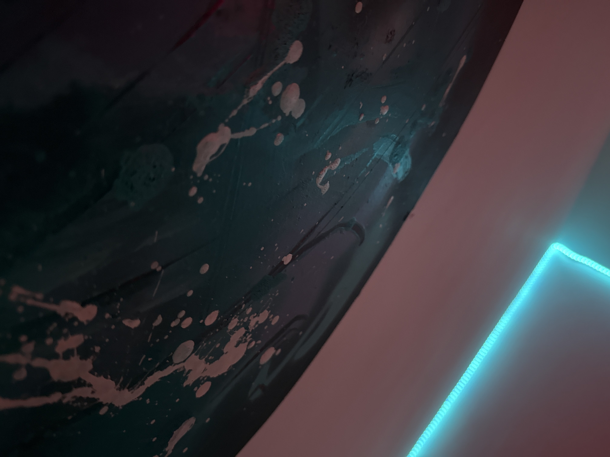 The image shows a close-up, abstract view of a surface with paint splatters. A bright blue neon light runs diagonally across the upper left portion of the frame, illuminating a pale pink wall or surface. The main subject is a dark, curved surface with numerous white and pinkish paint splatters of varying sizes. The splatters appear to be irregular and some are elongated, suggesting movement or dripping. The lighting on this surface is a mix of ambient light and a cool, teal-toned illumination, possibly from another light source not visible in the frame. There are subtle reflections and textures on the dark surface, indicating it might be glossy or metallic. The overall impression is one of a modern art piece or a creative process captured in a dimly lit environment. This scene is not indicative of a specific location in the city of Varna, Bulgaria, or any other identifiable geographical location.