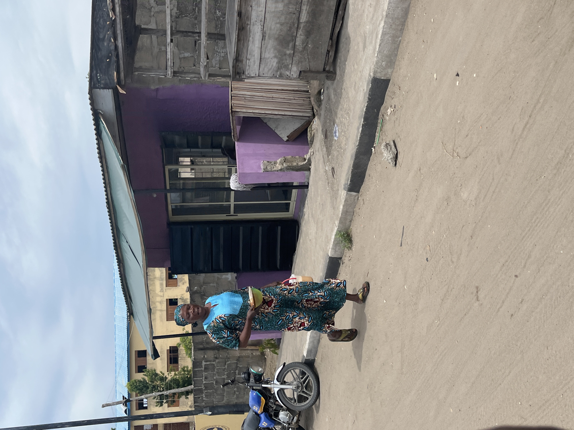 A woman in Badagry, Nigeria walks along a paved street, carrying items in her hands. She wears a blue and green patterned dress with a blue headscarf. A parked blue and yellow motorcycle is partially visible in the lower left of the image. Behind her, a purple building with a silver awning is seen. The street is adjacent to the building, with a concrete curb. The sky is overcast.