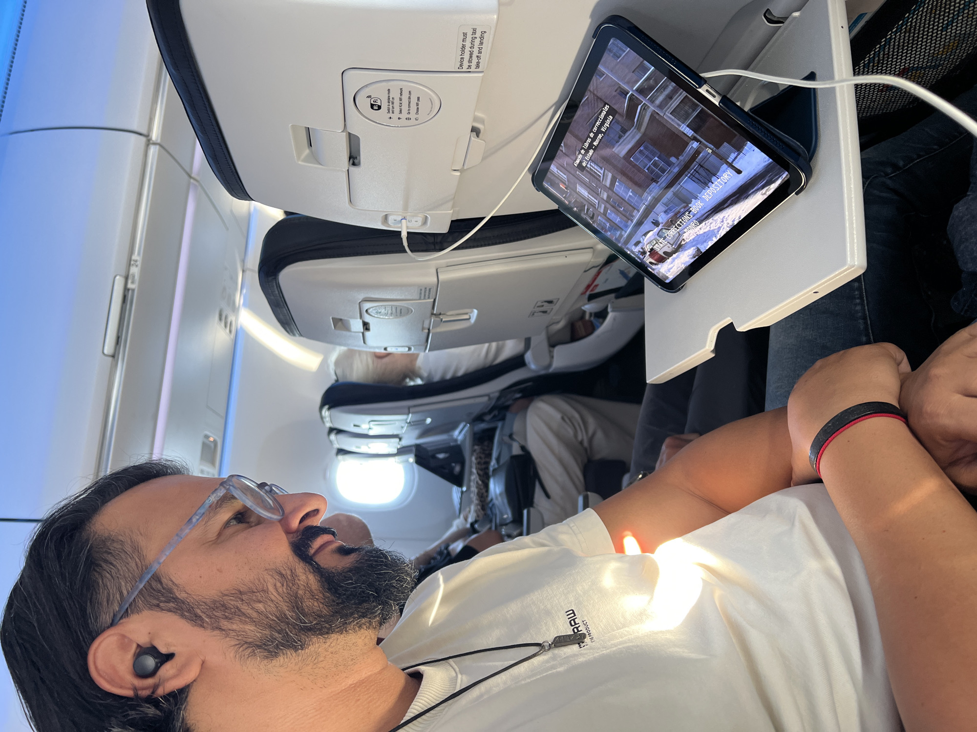 The image depicts a man sitting on an airplane, viewed from the chest up, wearing eyeglasses and wireless earbuds, and looking at a tablet. The tablet, propped on a foldable tray table, displays text and an image. The man is wearing a white t-shirt with a pendant necklace and a bracelet. The airplane cabin is visible, with seats and overhead compartments. The overall lighting suggests it's daytime, with sunlight entering the cabin. The provided crops offer closer views of the man's face, the airplane interior, and the tablet screen. The context provided by the user indicates the image is related to the city of Vijfhuizen, Netherlands.