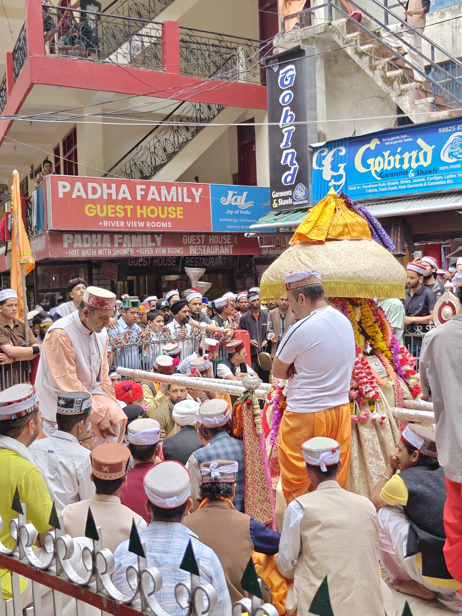 This image depicts a vibrant street scene in Kasol, India, likely during a religious procession or festival. At the center, a richly adorned palanquin or shrine, featuring abundant red, yellow, and purple flowers, garlands, and colorful fabrics, is topped with an ornate gold and yellow fabric umbrella. A man in a white t-shirt and orange dhoti stands with his back to the camera, attending to the central object.
Numerous men, many wearing traditional Himachali (Kullu) caps, are gathered around the palanquin, engaged in the event. To the left, a distinguished-looking man in a light orange kurta, white vest, and a beaded necklace is seated on a structure, possibly a religious elder or priest. Other spectators are visible behind a decorative metal railing in the foreground and lining the street, with some observing from the balconies of surrounding buildings.
The background features multi-story buildings, characteristic of a mountain town. Prominent red signage indicates "PADHA FAMILY GUEST HOUSE," advertising "RIVER VIEW ROOMS," "HOT BATH WITH SHOWER," and a "RESTAURANT." A vertical blue and black sign reads "GOBIIND Garments & Shawls," with a larger horizontal blue sign confirming "Gobind Garments & Shawls 100% PURE," which "Deals In:-Handloom Kullu Shawls, Stoles, Jackets, Cardigan, Ladies Tops, T-Shirts, Track Pants, Kids Garments & Cosmetics Etc." Numerous electrical wires crisscross overhead. The bright lighting suggests it is midday or early afternoon, with a lively yet respectful atmosphere among the participants.
