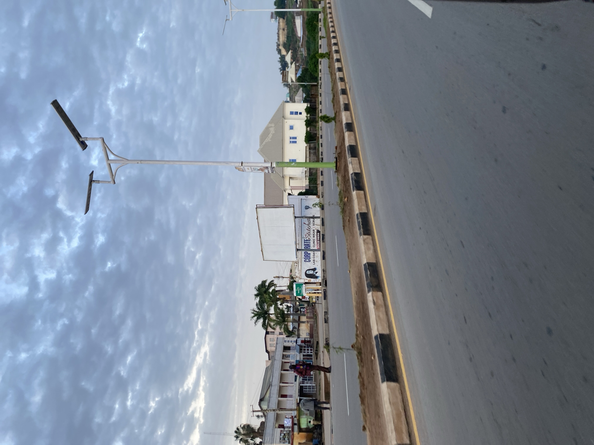 An eye-level, medium shot captures a street scene in Jos, Nigeria, under a cloudy sky. The foreground is dominated by a wide, grey asphalt road, with a yellow line marking the lane on the right and a concrete barrier with black and white diagonal markings separating the road from the earth-colored shoulder on the left. A few scattered dark spots are visible on the road surface.
In the mid-ground, a street lamp with two solar panels stands tall on a white pole. Beyond it, several buildings line the street. To the left, a two-story building with white and grey facades and balconies is visible, with palm trees in front. Two men are standing on the sidewalk in front of this building, one in a red and black plaid shirt and the other in a grey hooded jacket. Further down the street, a large white billboard is positioned. To the right of the billboard, a modern, light-colored house with a grey roof and blue-framed windows is situated, with smaller buildings and houses visible on a hill in the background. The landscape is somewhat hilly, with scattered trees and vegetation.
The overall impression is a typical urban environment in Jos, Nigeria, likely captured during the early morning or late afternoon due to the diffused light and cloud cover.