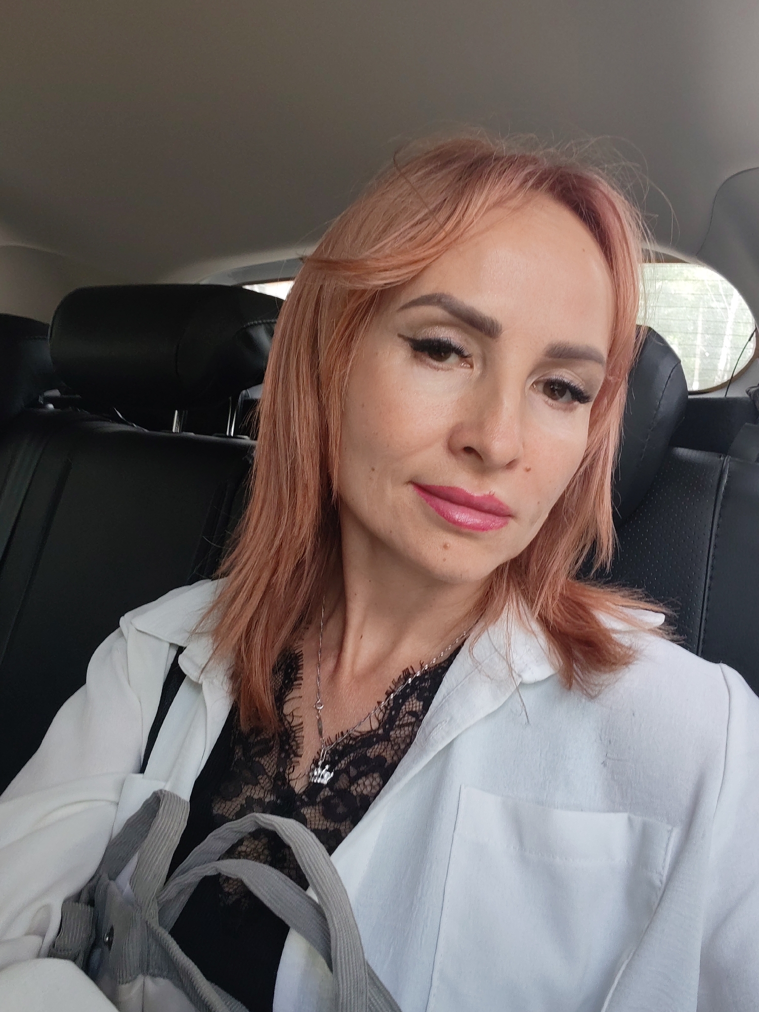 In the interior of a vehicle, a woman with peach-colored hair and a white blazer over a black lace top is captured in a selfie. She is wearing makeup and a necklace with a pendant. A gray bag is visible in the foreground. The background consists of car seats and a window, suggesting a daytime setting. The specific location of Chaikovsky, Russia, is not determinable from the visual information provided.