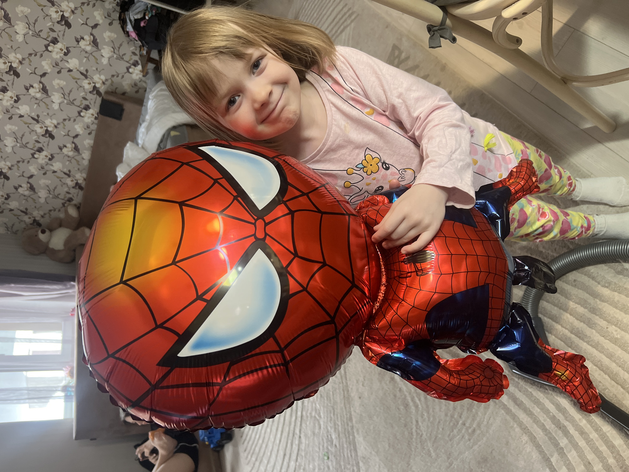 A young girl in pajamas is lying on a rug, hugging a large Spider-Man balloon. She has short blonde hair, fair skin, and appears to have rosy cheeks. Her pajamas are light pink with a giraffe-like cartoon on the shirt. The balloon is red with black web patterns and white eye patches. The background includes a patterned rug, a bed with clothing, a vacuum cleaner, and the legs of a wooden chair. The location is indicated as Chaikovsky, Russia.