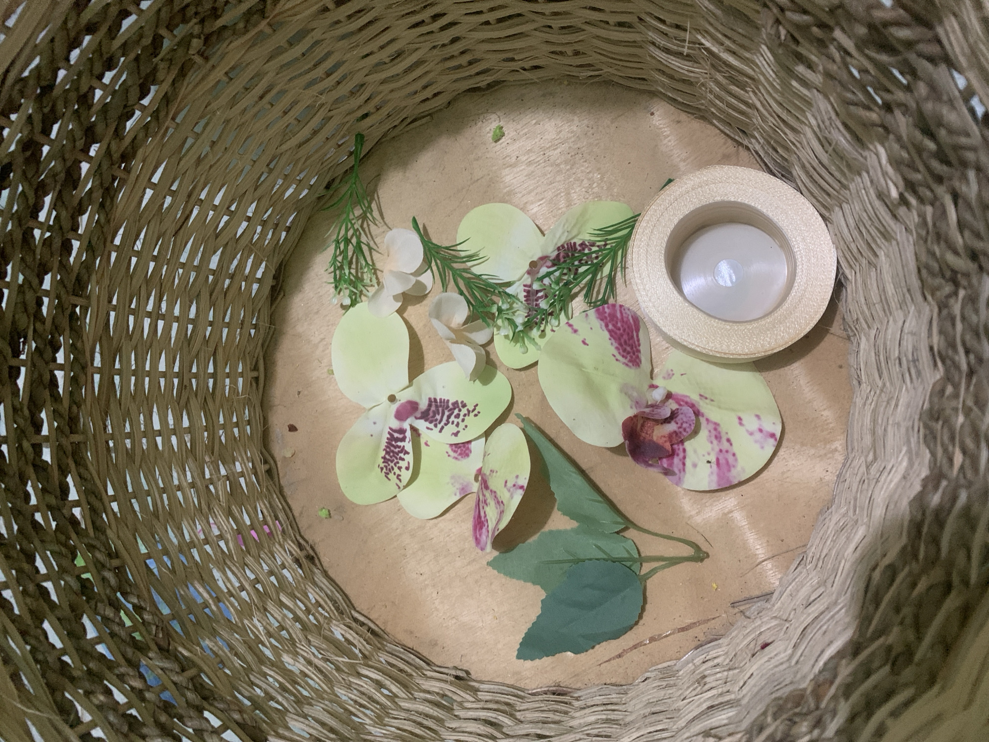 The image shows a close-up, top-down view of artificial flower petals and sprigs of greenery scattered inside a woven basket. A spool of pale gold ribbon with a white center is also visible. The basket appears to be made of natural fibers and has a textured, woven pattern. The petals are predominantly pale yellow with pink and purple splotches, suggesting they are part of an orchid design. The greenery consists of small, dark green leaves and delicate, needle-like sprigs. The background inside the basket is a light brown, wooden surface. The lighting suggests an indoor environment. There are no people or animals visible in the image. The scene is static, with no actions or interactions depicted. The location within the city of Kuala Pembuang, Indonesia, is not discernible from the visual information provided.