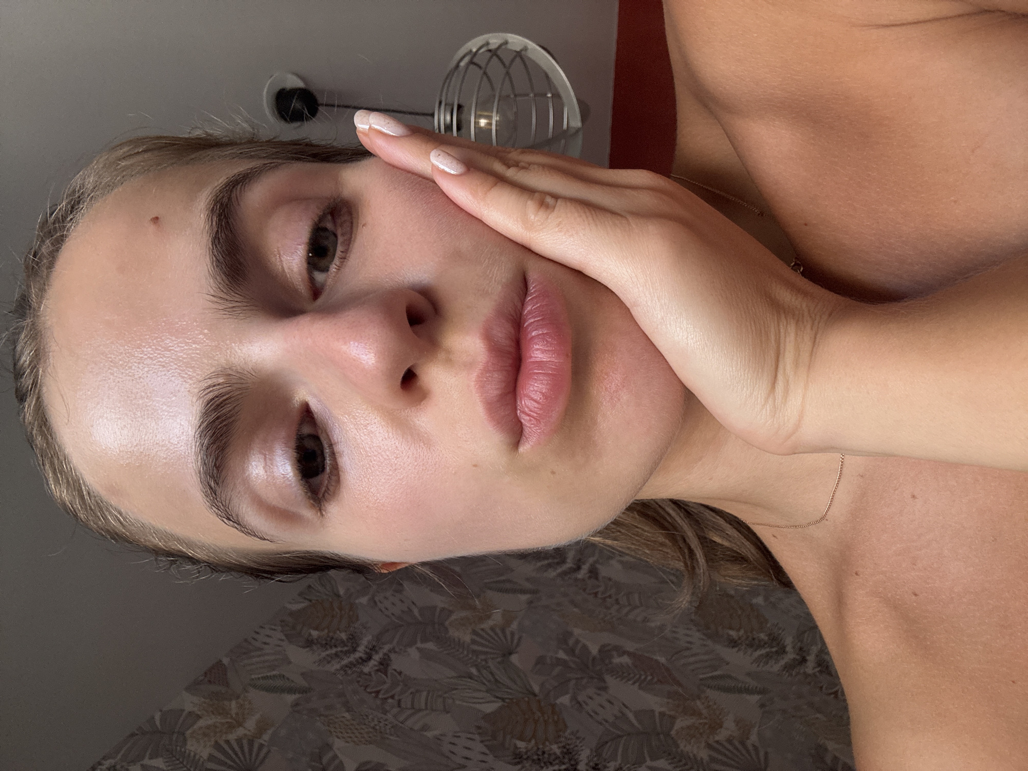 A close-up, head-and-shoulders selfie shows a young woman with her right hand resting on her cheek. Her lips are pursed. The background includes a patterned wallpaper on the left and a light fixture on the right. The woman is looking at the camera, and her skin has a dewy appearance. The lighting is soft, creating subtle highlights on her face. The image does not contain any information that would place it in the city of Vienne, France.
