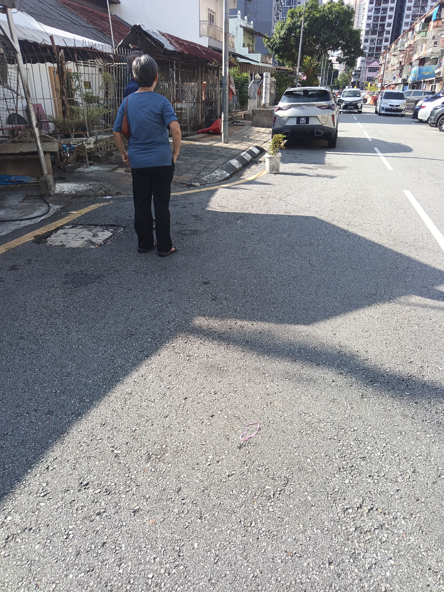 The image captures a street scene in Kuala Lumpur, Malaysia, likely a residential or commercial area with buildings lining the road. In the foreground, an individual, appearing to be an older woman with gray hair, stands facing away from the camera, dressed in a blue t-shirt and black trousers. She carries a brown handbag over her left shoulder. Her hands are in her pockets. Behind her, a modern silver SUV is parked on the right side of the road, facing away from the camera, with its license plate visible. Other vehicles are also present on the street, including a white sedan further down the road. The environment appears to be a typical urban street with asphalt pavement, a yellow line marking the road edge, and a sidewalk with some vegetation and debris. The lighting suggests it is daytime, with strong shadows cast by the buildings and vehicles. The buildings in the background display a mix of architectural styles, with some appearing older with corrugated metal roofs and others being modern high-rises. The scene conveys a sense of everyday life in an urban setting.