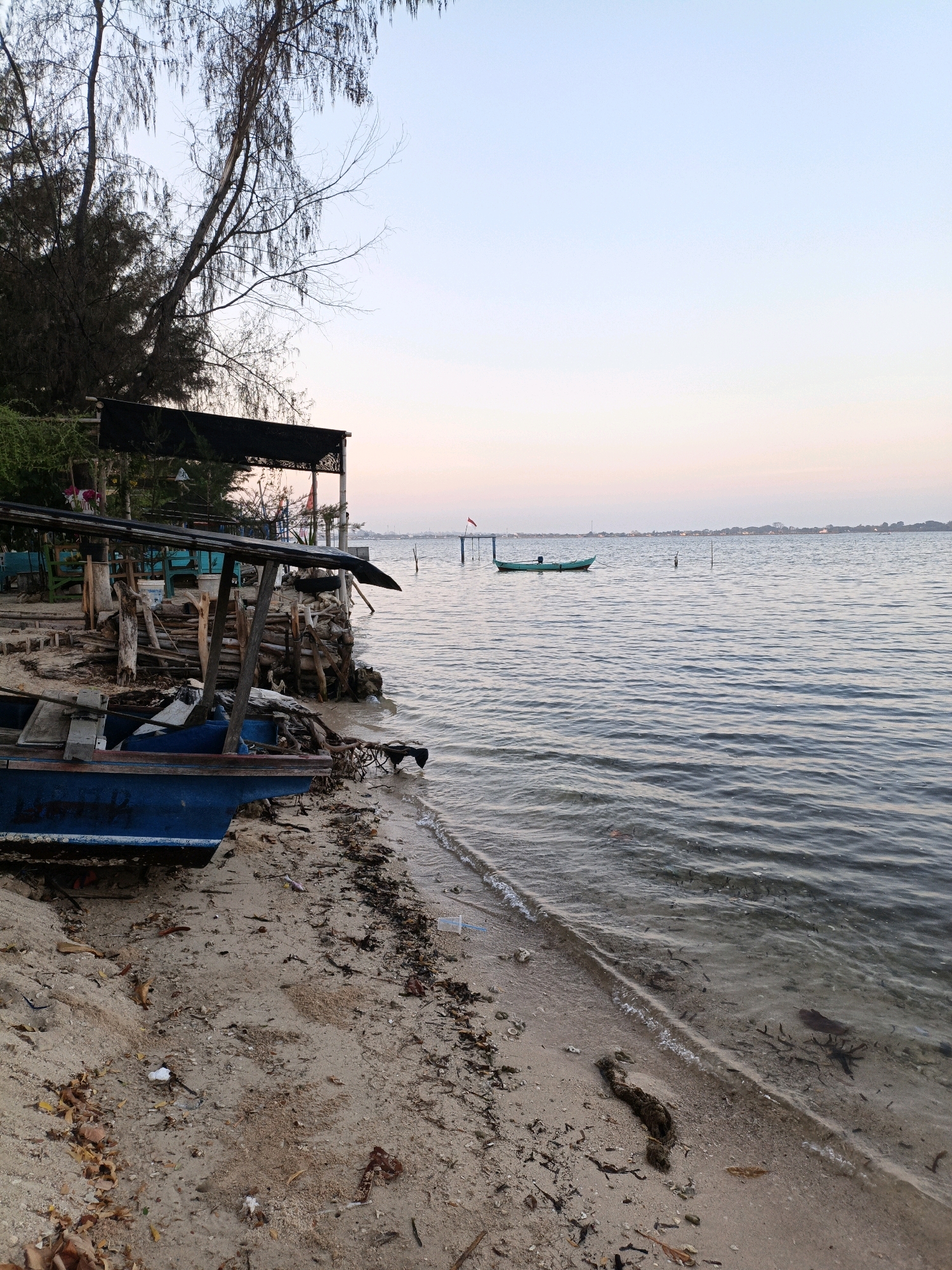 A tranquil scene unfolds at the Cikal, Indonesia shoreline, captured during the gentle light of dusk. The water, a serene expanse of rippling blues and grays, laps softly at the sandy beach. A lone, turquoise boat drifts peacefully in the distance, anchored by thin poles and marked by a small red flag. On the shore, a weathered blue boat rests partially on the sand, its hull showing signs of age and use. Behind it, a rustic wooden structure, likely a small pier or shelter, stands supported by weathered posts and planks. Debris, including dried leaves, plastic fragments, and driftwood, is scattered along the waterline, testament to the natural ebb and flow of the environment. The sky above transitions from a pale, soft blue to a delicate blush of pink and orange, indicating the day's end. The distant horizon is dotted with faint silhouettes of buildings and trees, hinting at a nearby settlement. The overall atmosphere is one of quietude and simple coastal life.