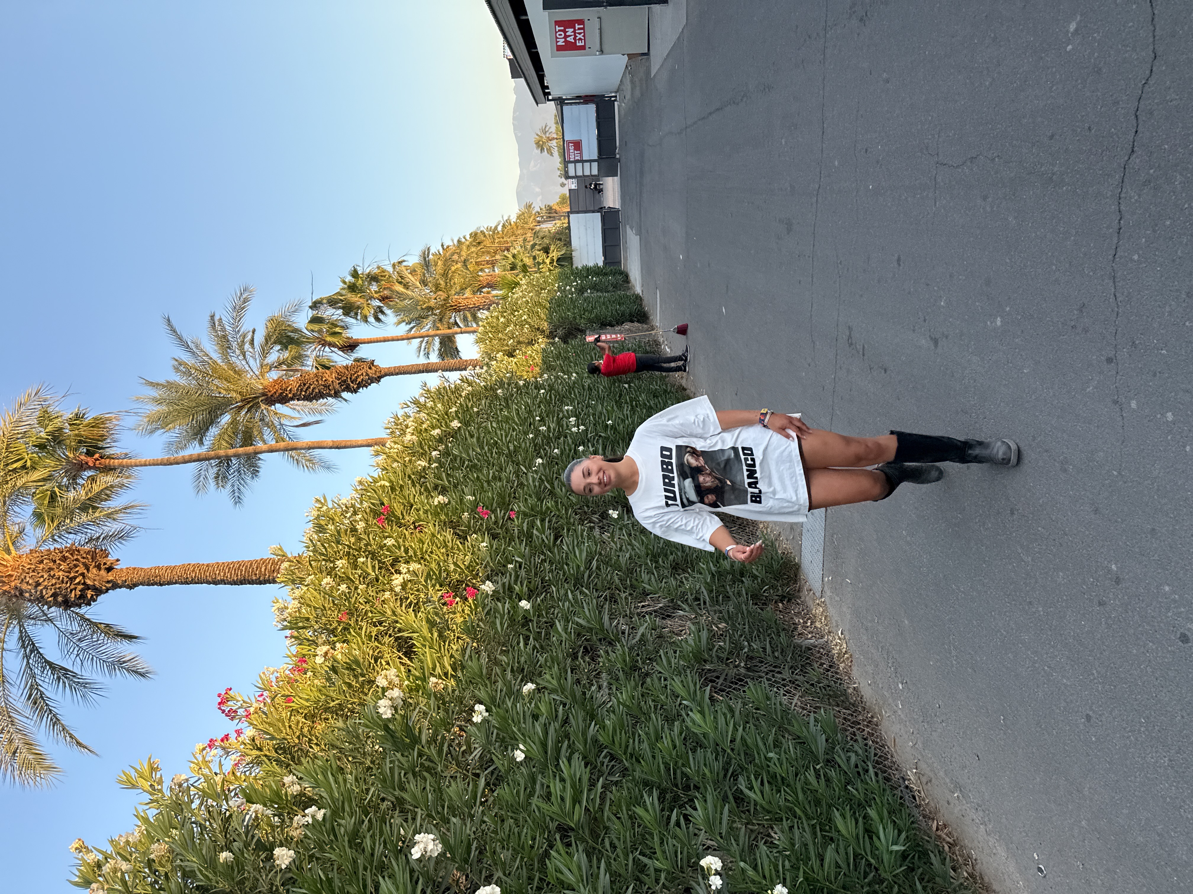 A woman in a white t-shirt and boots is walking along a paved road in Indio, United States. The t-shirt has an image of "TURBO BLANCO" printed on it. Palm trees line one side of the road. There is a long flowering bush on the left side of the road next to the palm trees. A man in a red shirt is sweeping in the background. A building labeled "NOT AN EXIT" is visible.