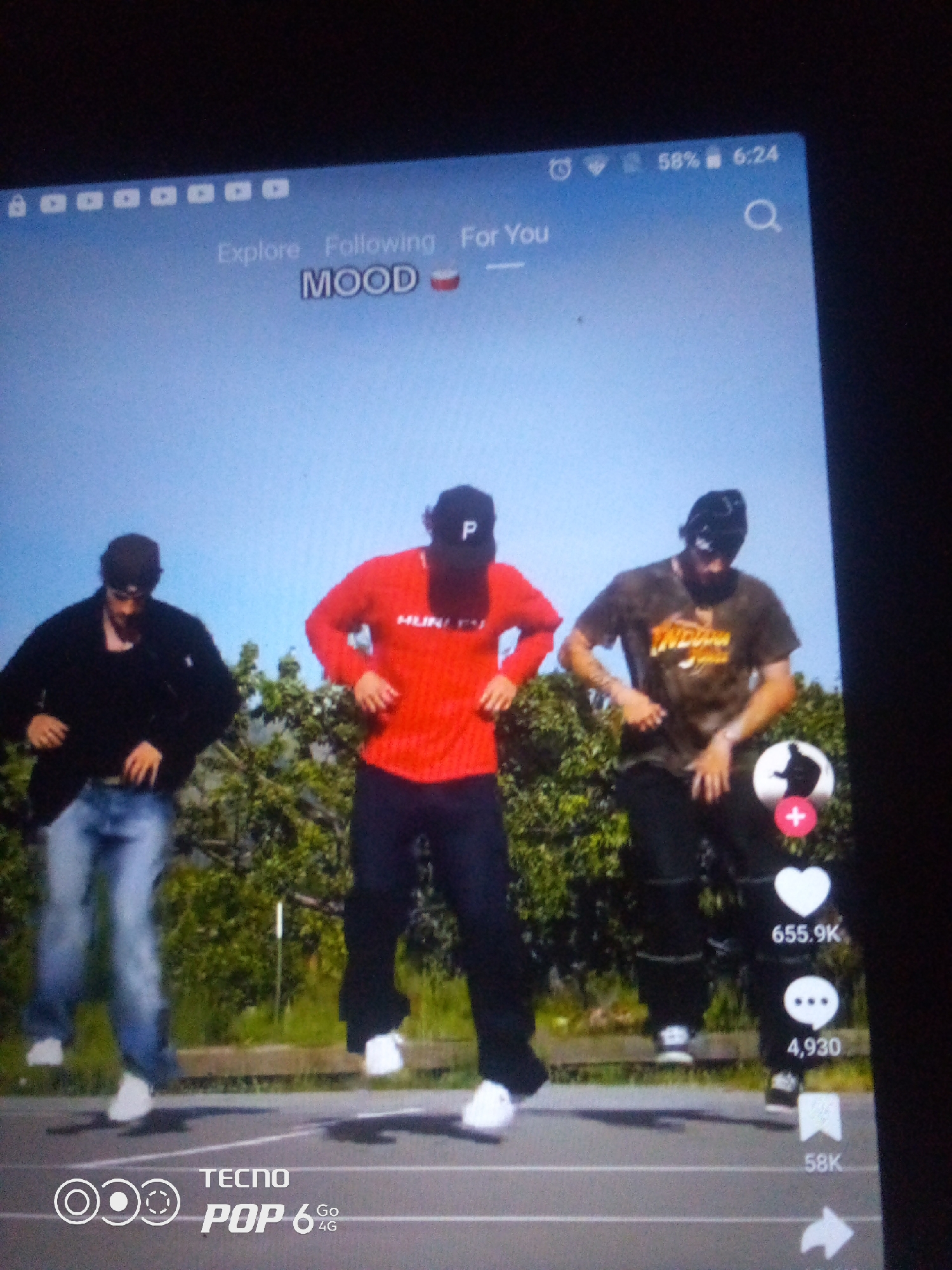 The image is a screenshot of a mobile device displaying a video on a social media platform. Three young men are captured mid-dance outdoors, with a clear blue sky and lush green foliage in the background. The individuals are dressed casually: one in a black jacket and blue jeans, another in a red long-sleeved shirt and dark pants, and the third in a dark, patterned t-shirt and black pants. They all appear to be wearing baseball caps.
The video interface shows interface elements common to social media apps, including icons for likes, comments, and shares with corresponding numerical values. Text at the top of the screen reads "Explore Following For You" and "MOOD" with an emoji. A battery indicator shows 58%, and the time is 6:24. The bottom of the screen displays "TECNO POP 6 Go 4G" branding.
There is no visible information within the image that would indicate the location as Howayi, Nigeria.