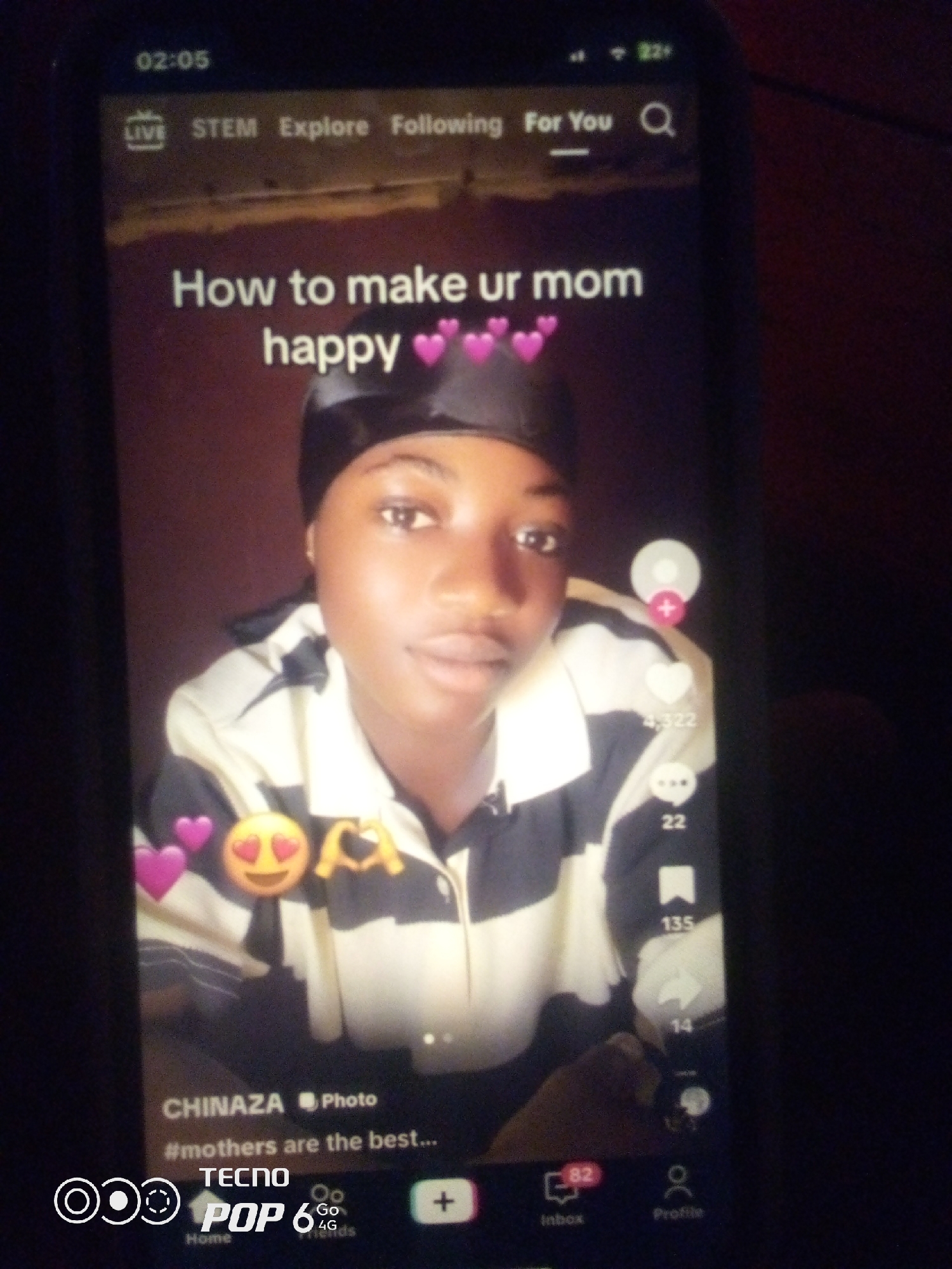 The image is a TikTok video featuring a young woman with a headscarf. The video's title reads "How to make ur mom happy" and includes pink heart emojis. The user's name is CHINAZA, and the caption is "#mothers are the best...". The bottom of the screen shows the TECNO POP 6 phone brand. The video has a like count of 4,322 and a comment count of 22. The overall environment appears to be indoors with a dark background. There is no discernible information about the location within the city of Howayi, Nigeria.