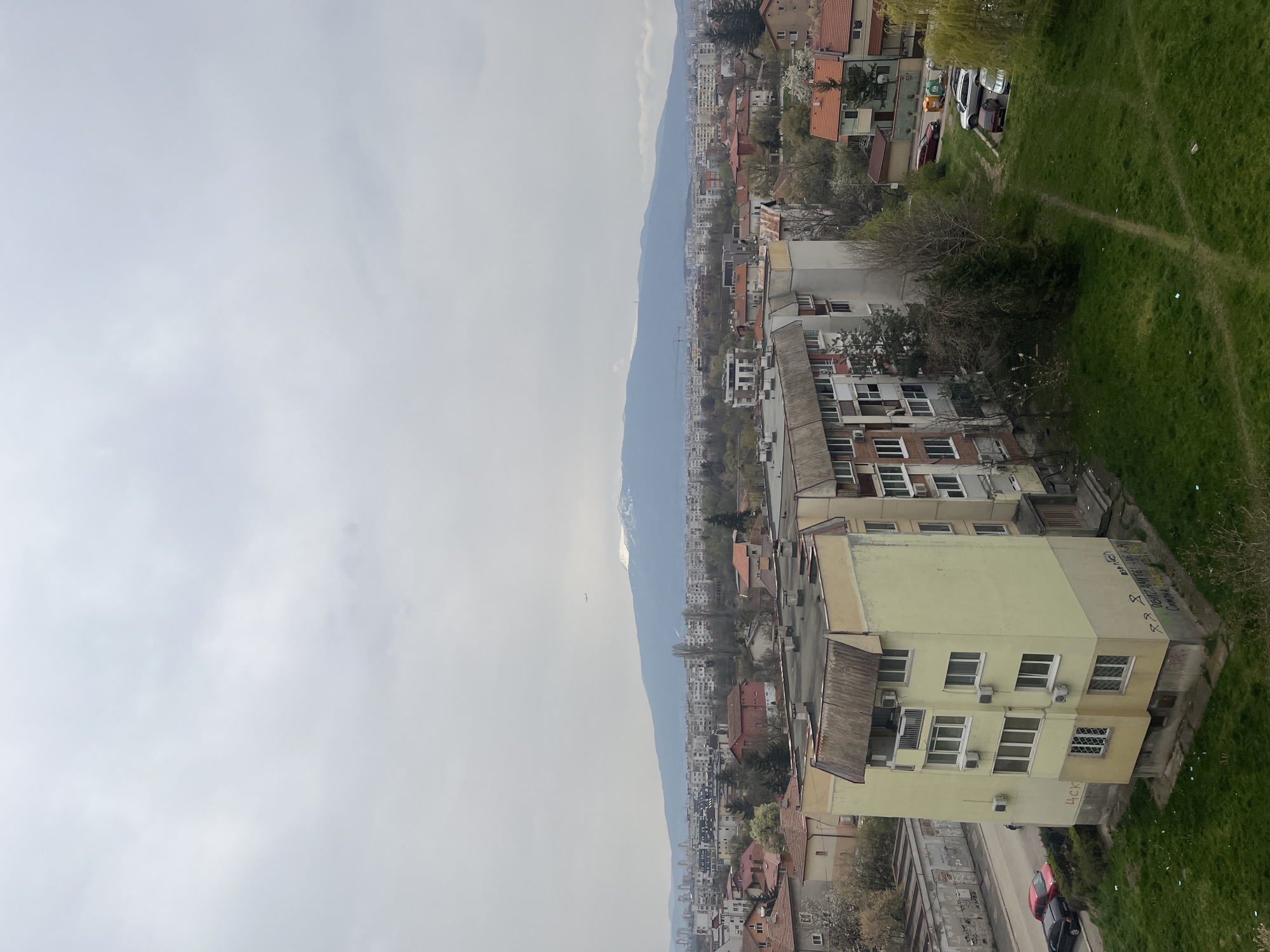 The image shows an elevated view of a cityscape in Sofia, Bulgaria, with a mountain range in the background. The sky is overcast with clouds. In the foreground, there are buildings of varying heights and architectural styles, with a mix of residential and commercial properties. A green grassy area is visible on the right side of the frame, potentially part of a park or a residential yard. The streets are visible, with some cars parked along the sides. The overall environment is urban, with a mix of older and more modern structures.