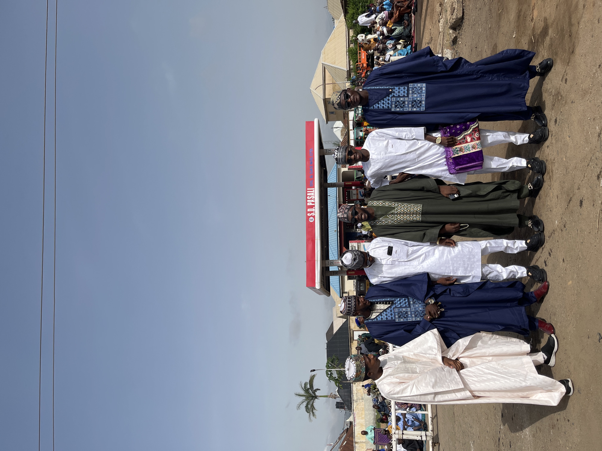 Six men stand in a line outside the S.D. Pasali filling station. The men are wearing traditional clothing, including hats, kaftans, and baban riga gowns. A crowd of people gathers in the background. The sky is blue. The scene takes place in Jos, Nigeria.