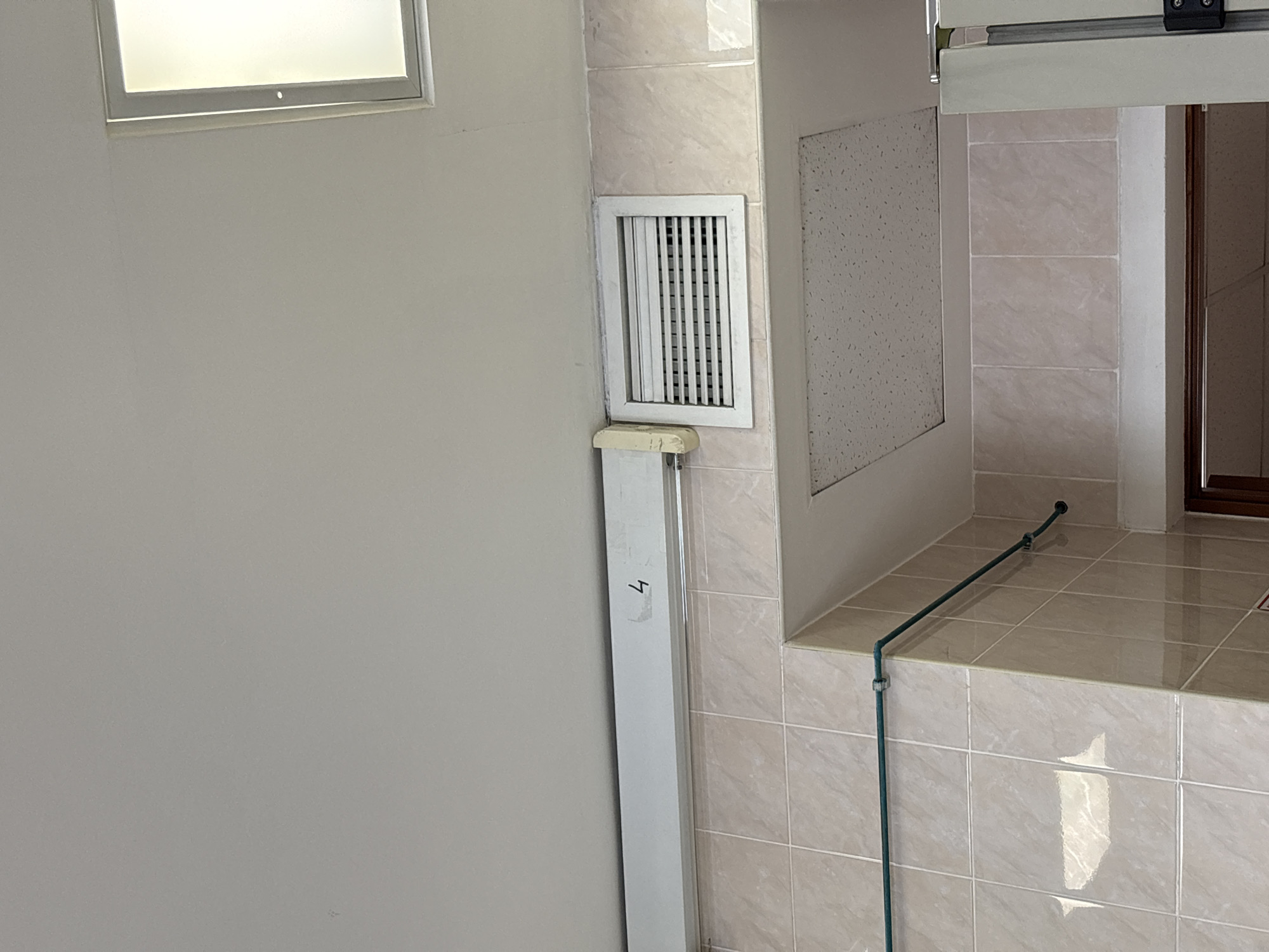 This interior image captures a utilitarian space, possibly a bathroom, utility room, or hallway in Saint Petersburg, Russia. The scene is devoid of people.
On the left, a plain, light-colored wall occupies most of the frame, with a rectangular window featuring frosted glass and a white frame visible in the upper left corner. The window suggests natural light illuminates the space.
To the right, the wall is covered with light beige or cream-colored square tiles, which extend onto a tiled countertop or shelf. A white rectangular ventilation grille is prominently placed on the tiled wall. Below the grille, a long, vertical white fixture, possibly a fluorescent light cover or a conduit, extends downwards. The number "4" is visible in black on this fixture.
Further to the right, a square, light-colored panel with a speckled texture is set into the wall above the tiled shelf, possibly an access panel or a ceiling tile. A dark green pipe or hose runs vertically along the tiled wall, bends at the tiled shelf, and continues horizontally across it, secured by small clips. A dark brown window or door frame is partially visible behind the tiled shelf in the far right background, with a white sliding track mechanism visible above it.
The overall impression is one of a functional, brightly lit interior space. The visible text is the number "4".