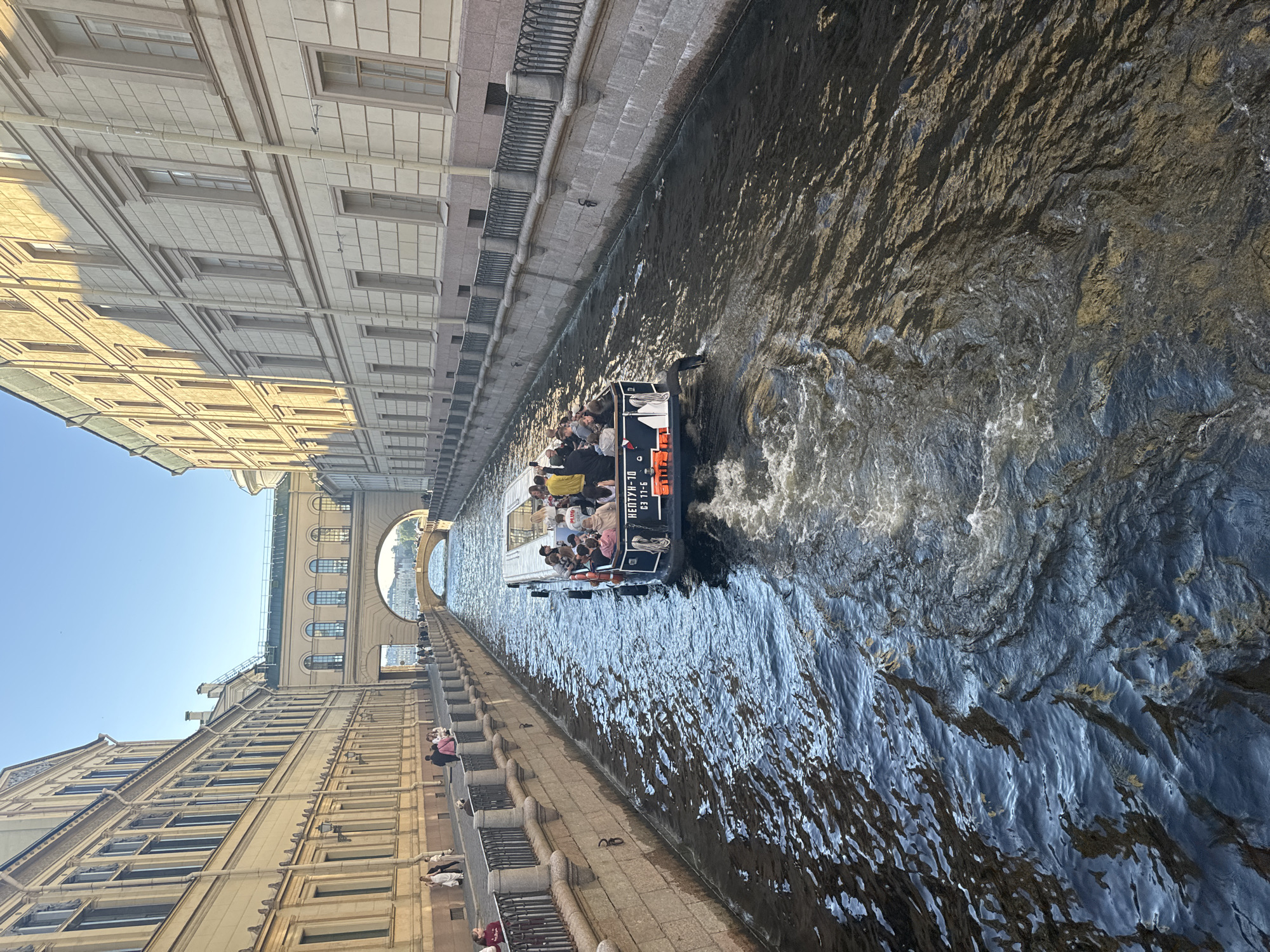 This image depicts a scenic canal in Saint Petersburg, Russia, under a clear blue sky, suggesting a pleasant day. The sun is casting shadows, indicating it is likely daytime, possibly late morning or afternoon. The waterway is lined with ornate, classical buildings, characterized by their light-colored facades, numerous windows, and detailed architectural elements. A stone embankment with railings runs along the side of the canal.
A tour boat filled with people is navigating the canal, creating ripples in the water. The passengers appear to be enjoying a leisurely tour, with some looking around and others conversing. The boat is dark and has life vests visible, suggesting it is designed for passenger excursions.
Visible text on the boat reads "НЕЙТУН-10" and "63 TT-5". This is likely the name of the boat and its registration number. The overall mood of the image is one of tranquil tourism and appreciation for the city's historic architecture and waterways. The perspective from which the photo is taken suggests the photographer is either on another boat or standing on a bridge, looking down the canal towards a distant arched bridge.