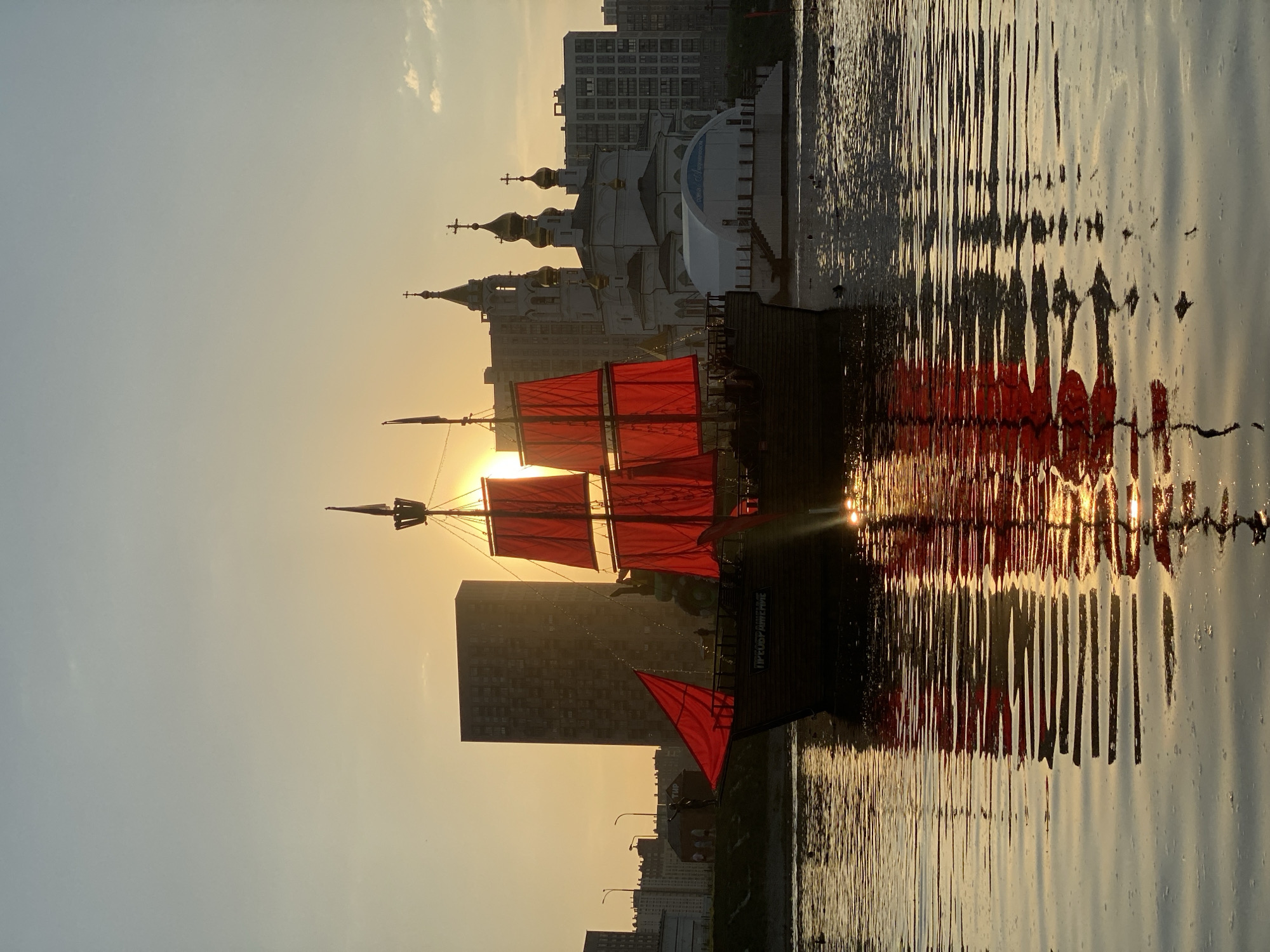 The image captures a scene in Yekaterinburg, Russia, featuring a large ship with red sails. The ship is docked or sailing on a body of water, likely a river or lake. Its reflection is visible in the water. A Russian Orthodox church with golden domes stands in the background, along with modern residential buildings. The image is taken at sunset or sunrise, casting a warm, golden light across the scene. The scene depicts the Peter's fleet of ships and a fragment of the landscape of Yekaterinburg.